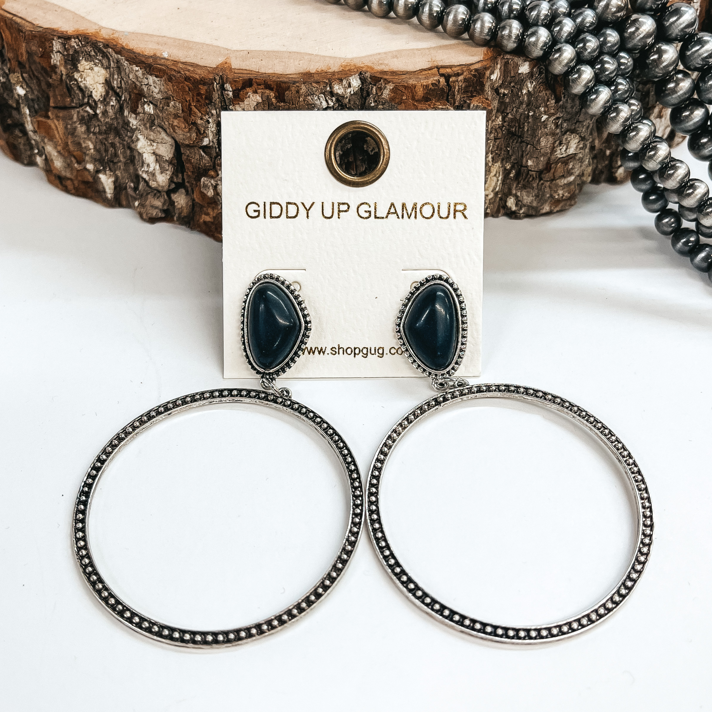 Asymmetrical stone stud earrings in black with a  silver, textured open circle pendant hanging from  the bottom. These earrings are  pictured on a white background, leaning up against a  slab of wood and withe faux navajo pearls in the back  as decor.