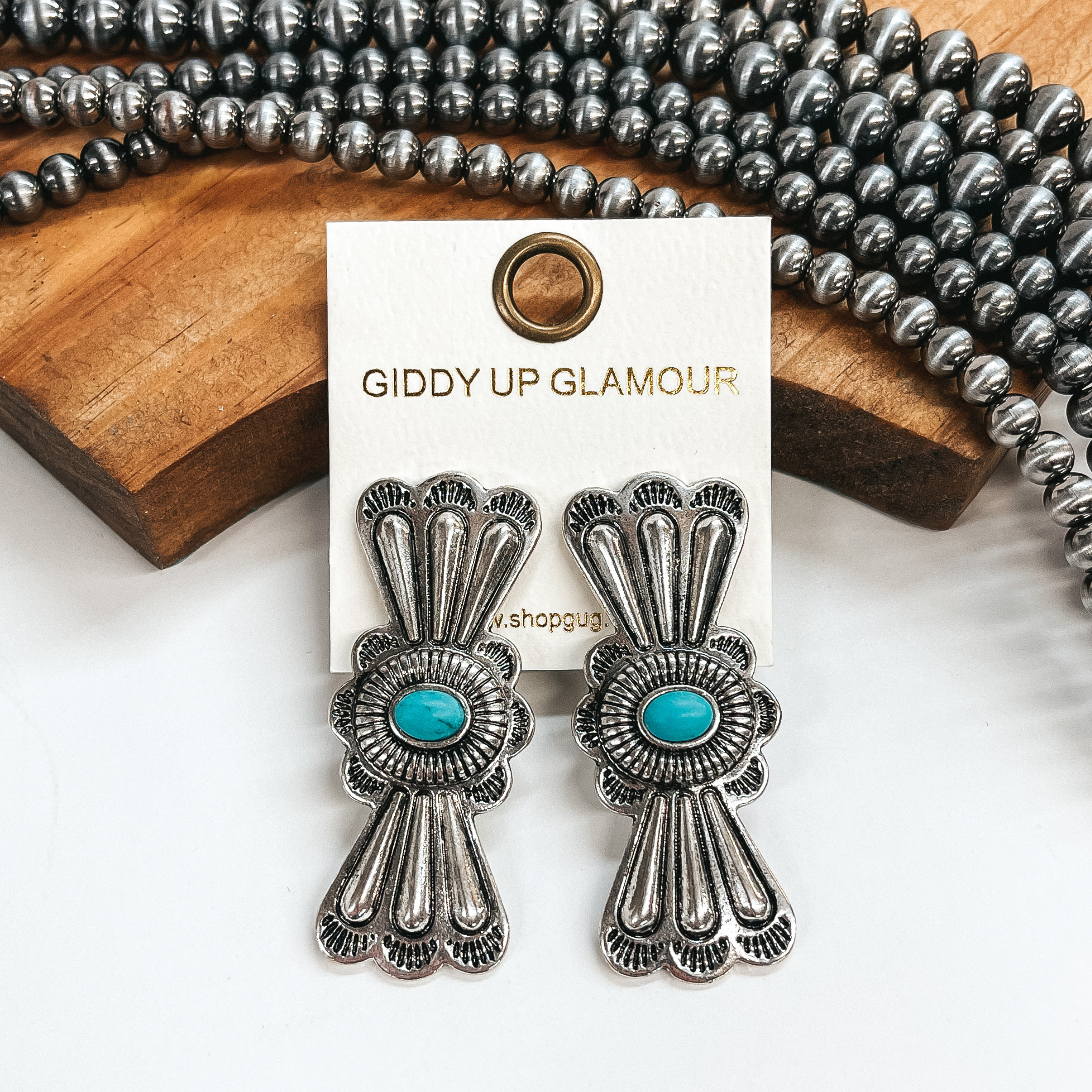 These are hour glass shaped earrings in silver with  a concho pattern. They have a small faux turquoise  stone in the center. These earrings are taken on a  white backround, leaned up against a brown block with  faux navajo pearls in the back as decor.