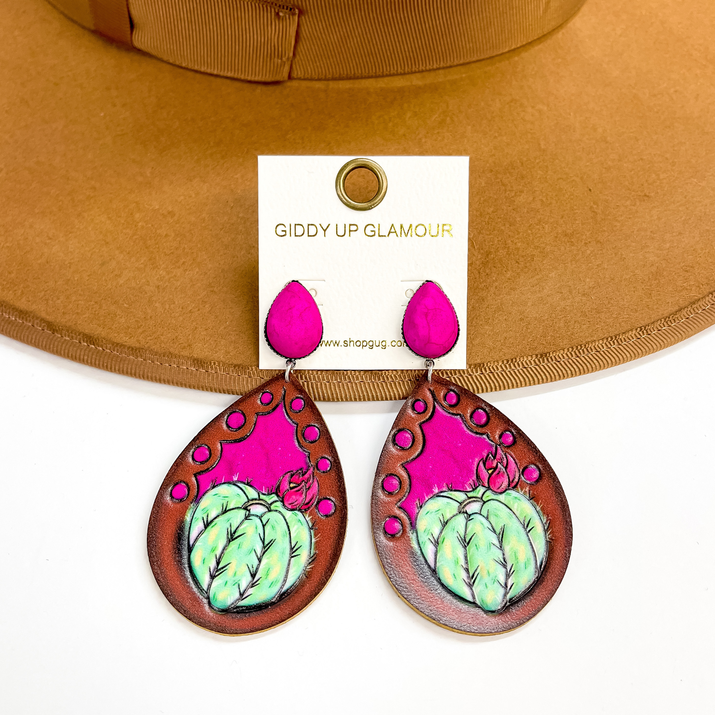These are hot pink teardrop stone earrings in a  silver setting with a brown leather teardrop. The teardrop has a small green catucs with a pink  flower in a hot pink background. These earrings are  taken on a brown hat brim and a white background.