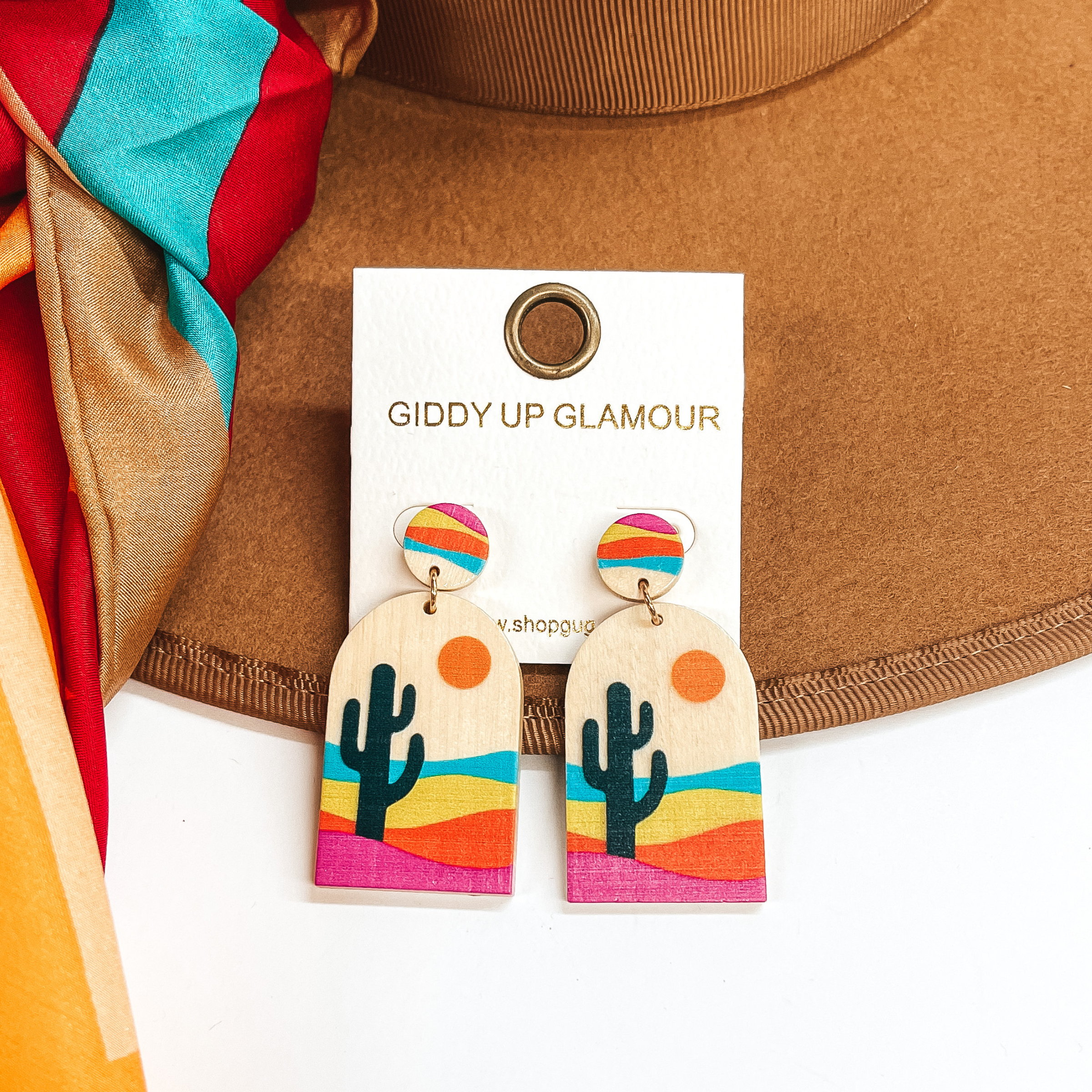 These are wood arch shape earrings with a circle  post back. These earrings has a dark green cactus  in a desert with turquoise, orange, yellow, and pink  colors. These earrings  are taken on a brown hat brim and a white background  with a multicolored wild rag in the side as decor.