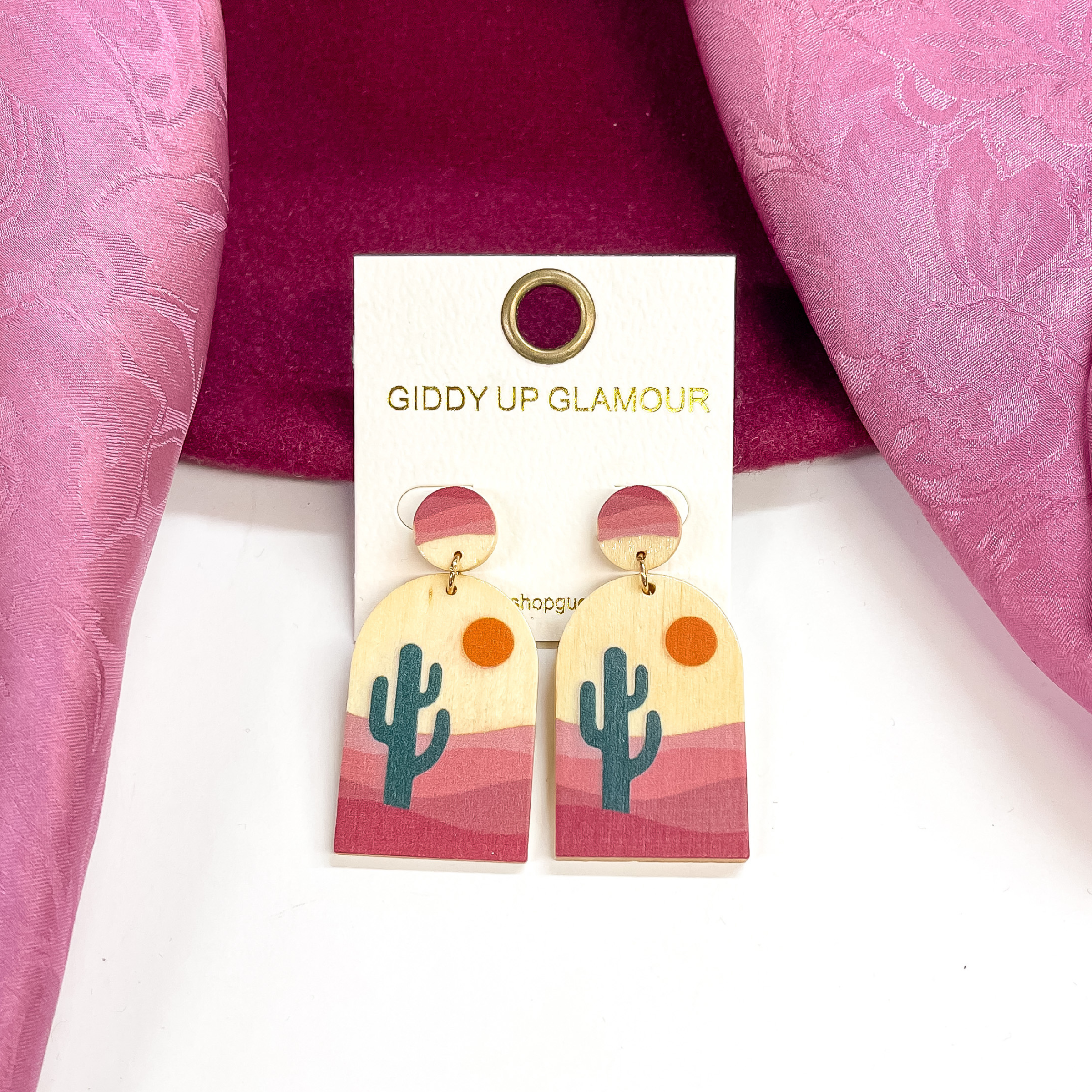 These are wood arch shape earrings with a circle  post back. These earrings has a dark green cactus  in a desert with pink mauve colors. These earrings  are taken on a burgundy hat brim and a white  background, with a light purple wild rag in the sides  as decor.