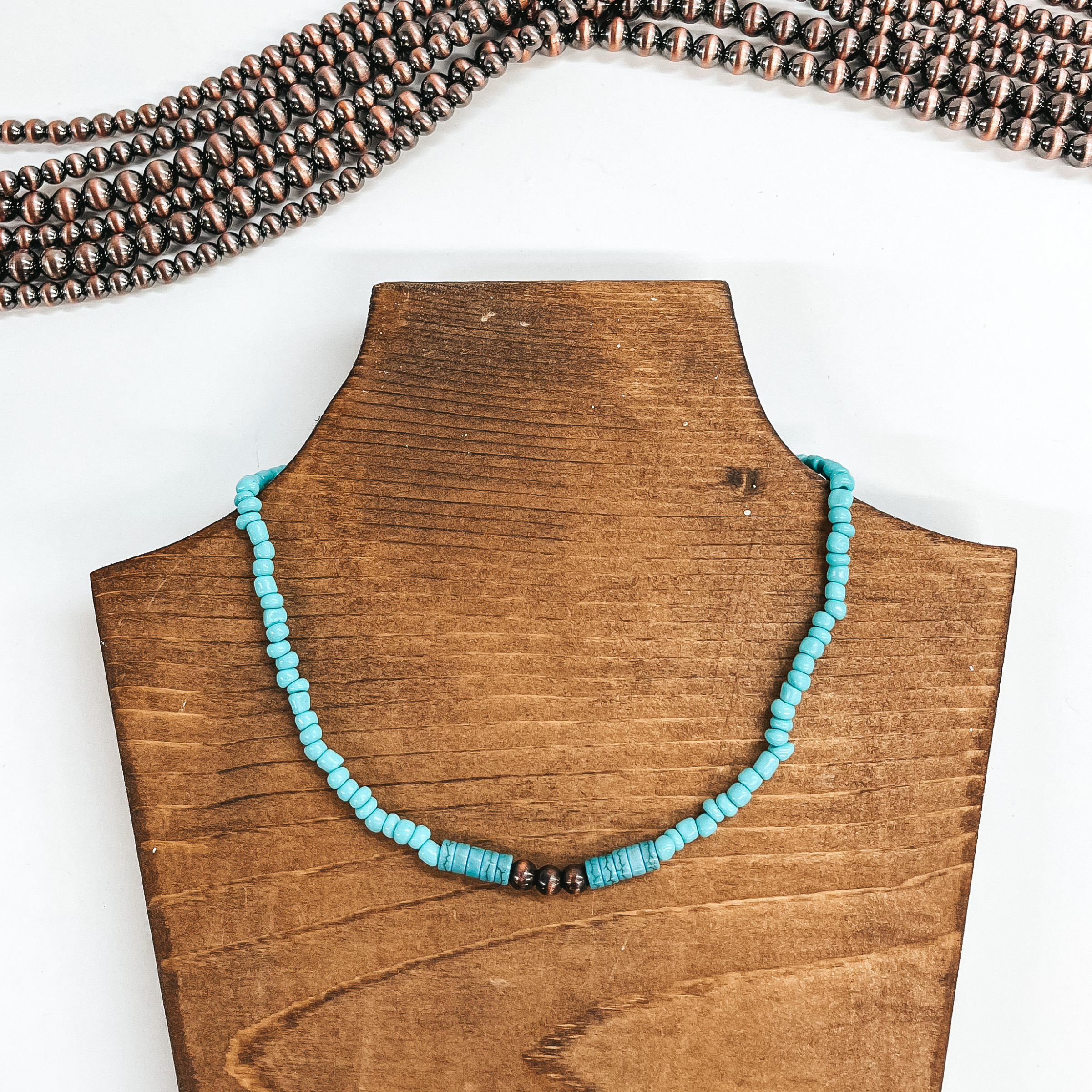 This is a turquoise beaded asymmetrical beads with  five teal/dark turquoise beads with three copper  faux navajo pearls in the center. This necklace is  taken on a brown necklace holder and white  background, their are copper navajo pearls in the  top as decor.