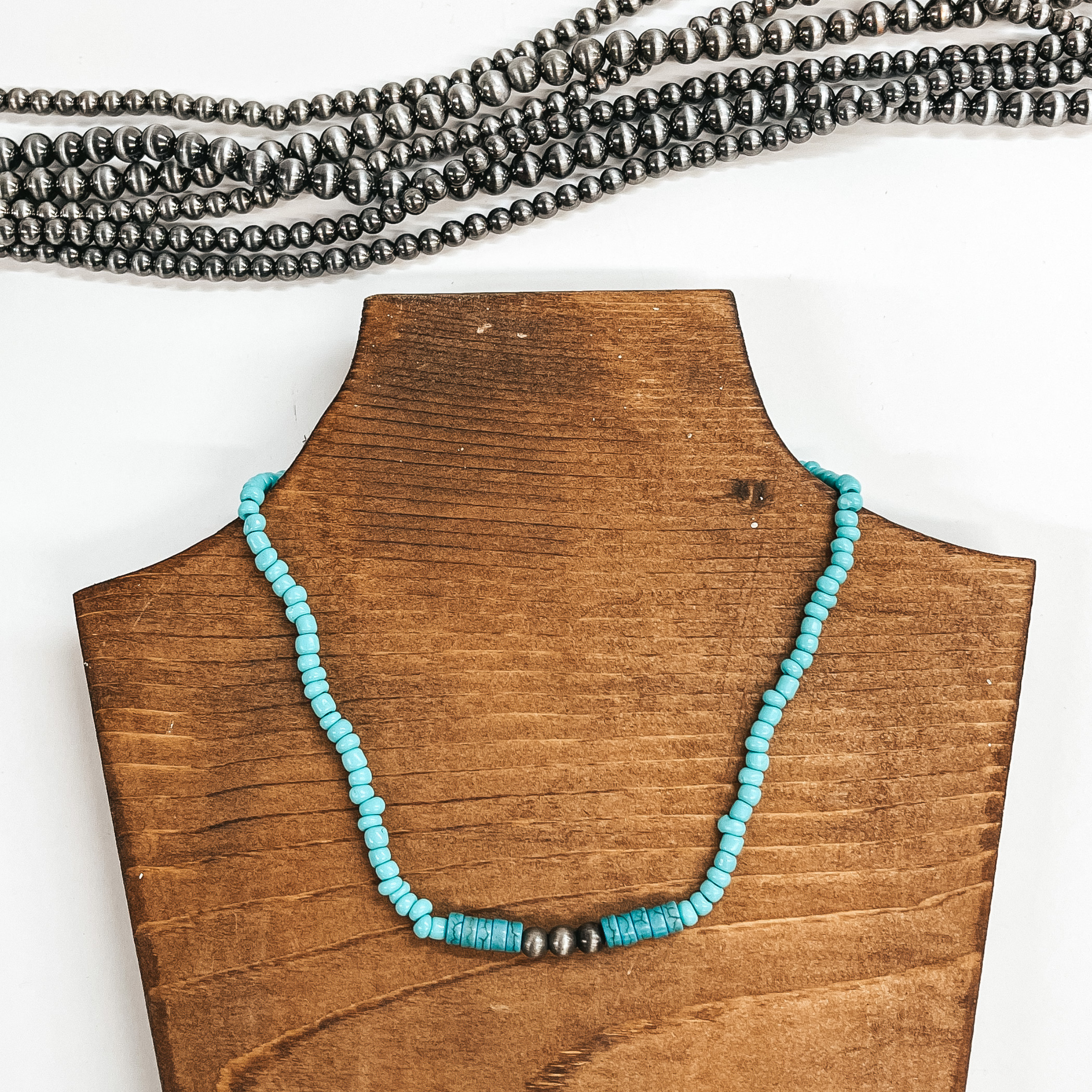 This is a turquoise beaded asymmetrical beads with  five teal/dark turquoise beads with three silver  faux navajo pearls in the center. This necklace is  taken on a brown necklace holder and white  background, their are silver navajo pearls in the  top as decor.