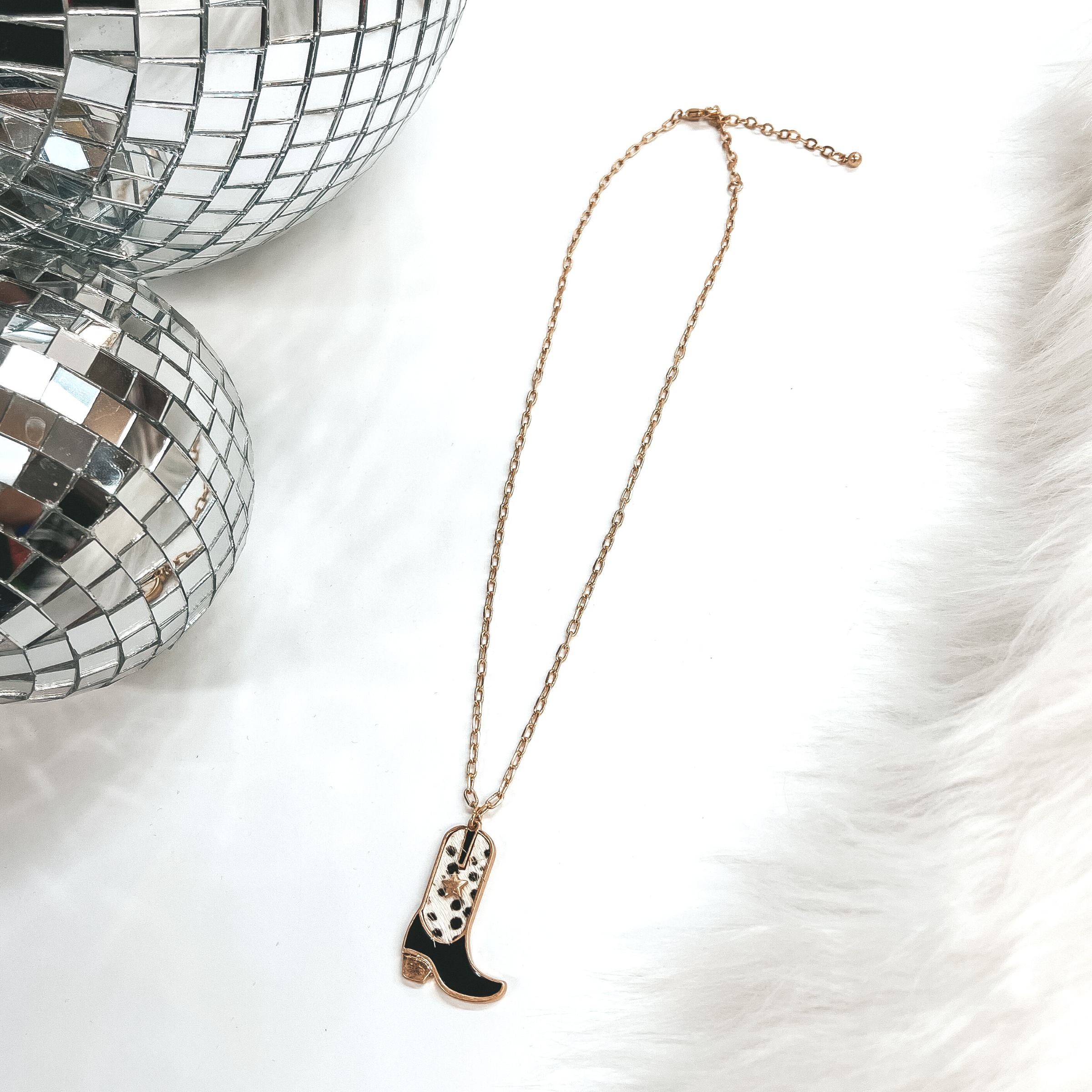 This is a gold chain necklace with a boot pendant  in a gold setting. The boot pendant is black and  has a dotted print with a gold star. This necklace  is taken on laying on a white background with a  white fur carpet in the side and two disco ball as  decor.