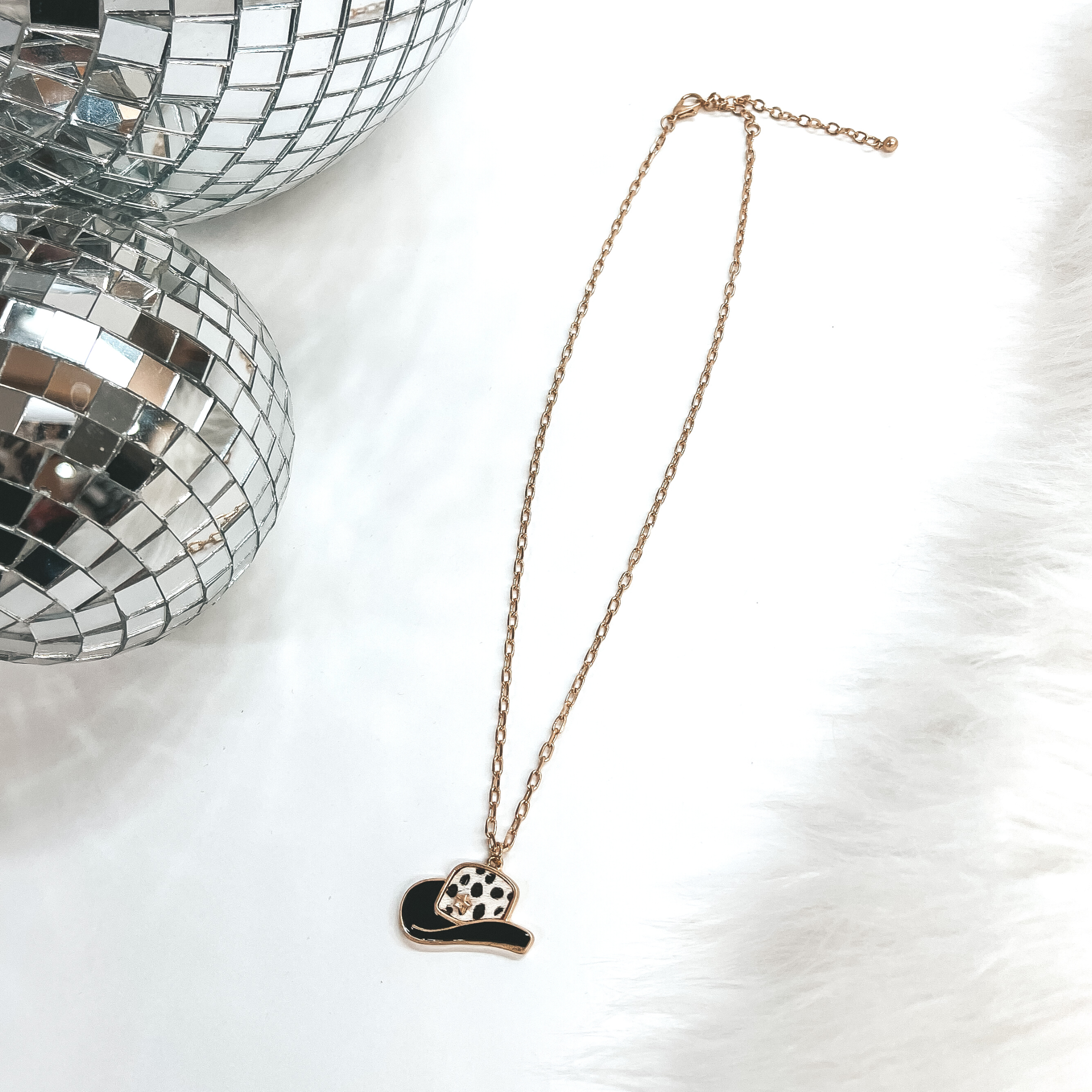 This is a gold chain necklace with a hat pendant in  a gold setting. The hat pendant is black and has  a dotted print with a gold star. This necklace is  taken on laying on a white background with a white  fur carpet in the side and two disco ball as decor.