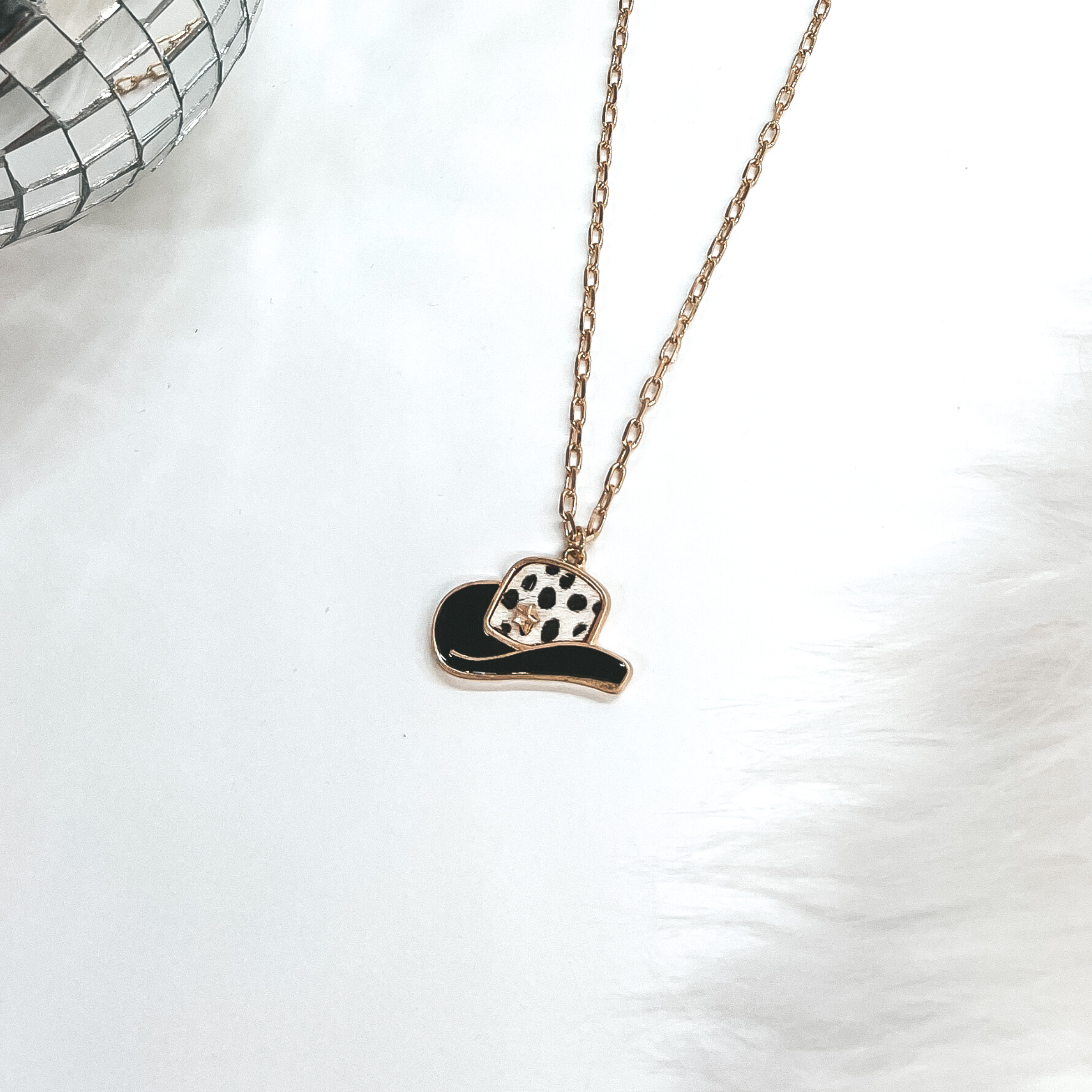 This is a gold chain necklace with a hat pendant in a gold setting. The hat pendant is black and has a dotted print with a gold star. This necklace is taken on laying on a white background with a white fur carpet in the side and two disco ball as decor.