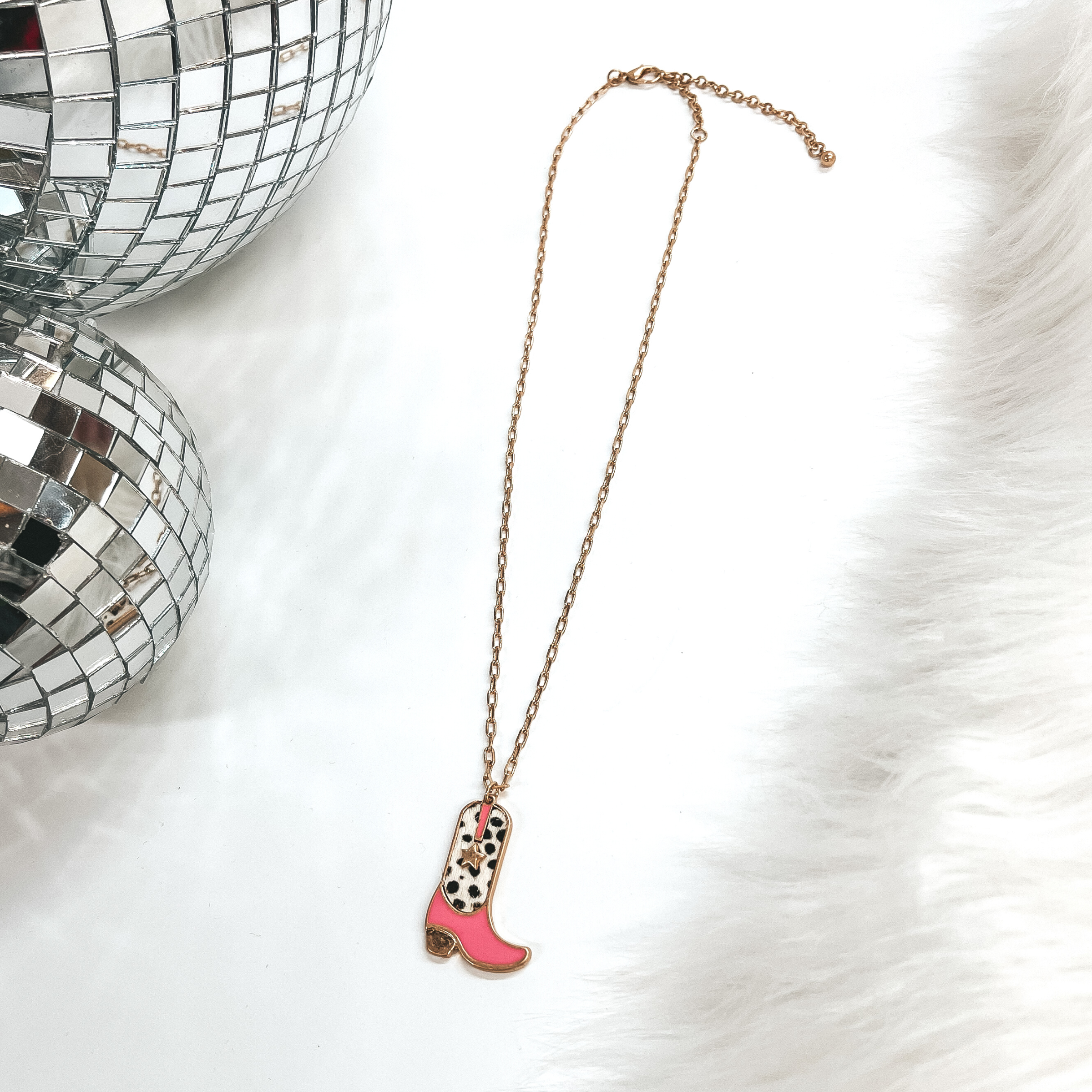 This is a gold chain necklace with a boot pendant  in a gold setting. The boot pendant is pink and  has a dotted print with a gold star. This necklace  is taken on laying on a white background with a  white fur carpet in the side and two disco ball as  decor.