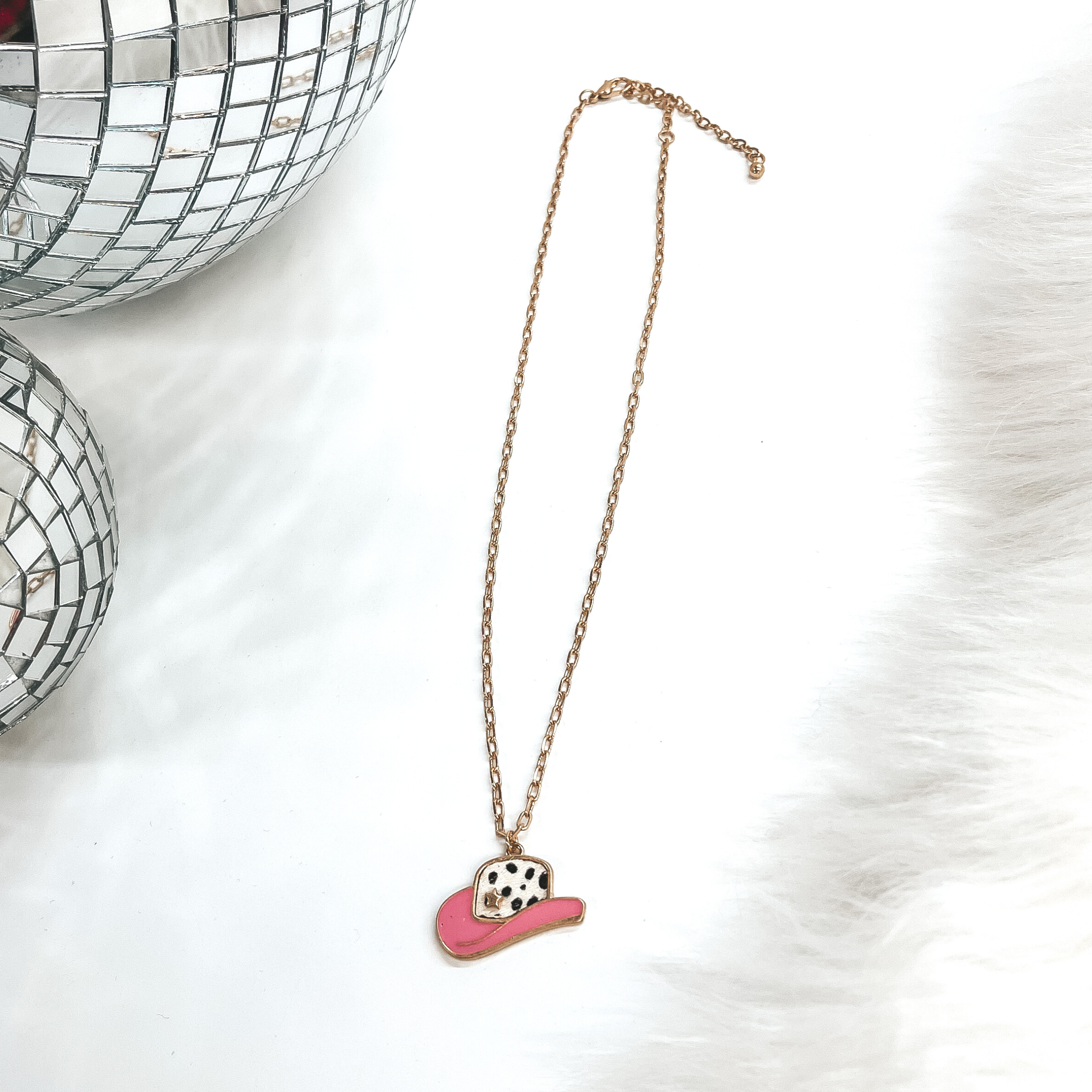 This is a gold chain necklace with a hat pendant in  a gold setting. The hat pendant is pink and has  a dotted print with a gold star. This necklace is  taken on laying on a white background with a white  fur carpet in the side and two disco ball as decor.