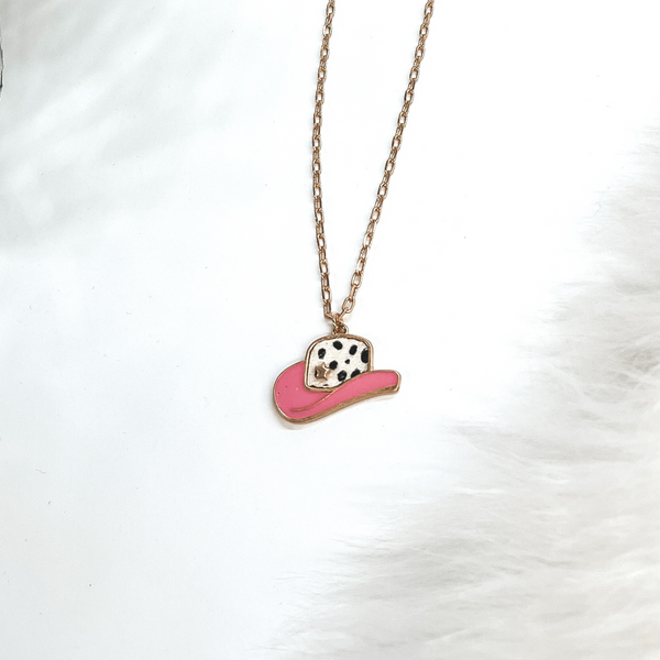 This is a gold chain necklace with a hat pendant in a gold setting. The hat pendant is pink and has a dotted print with a gold star. This necklace is taken on laying on a white background with a white fur carpet in the side and two disco ball as decor.