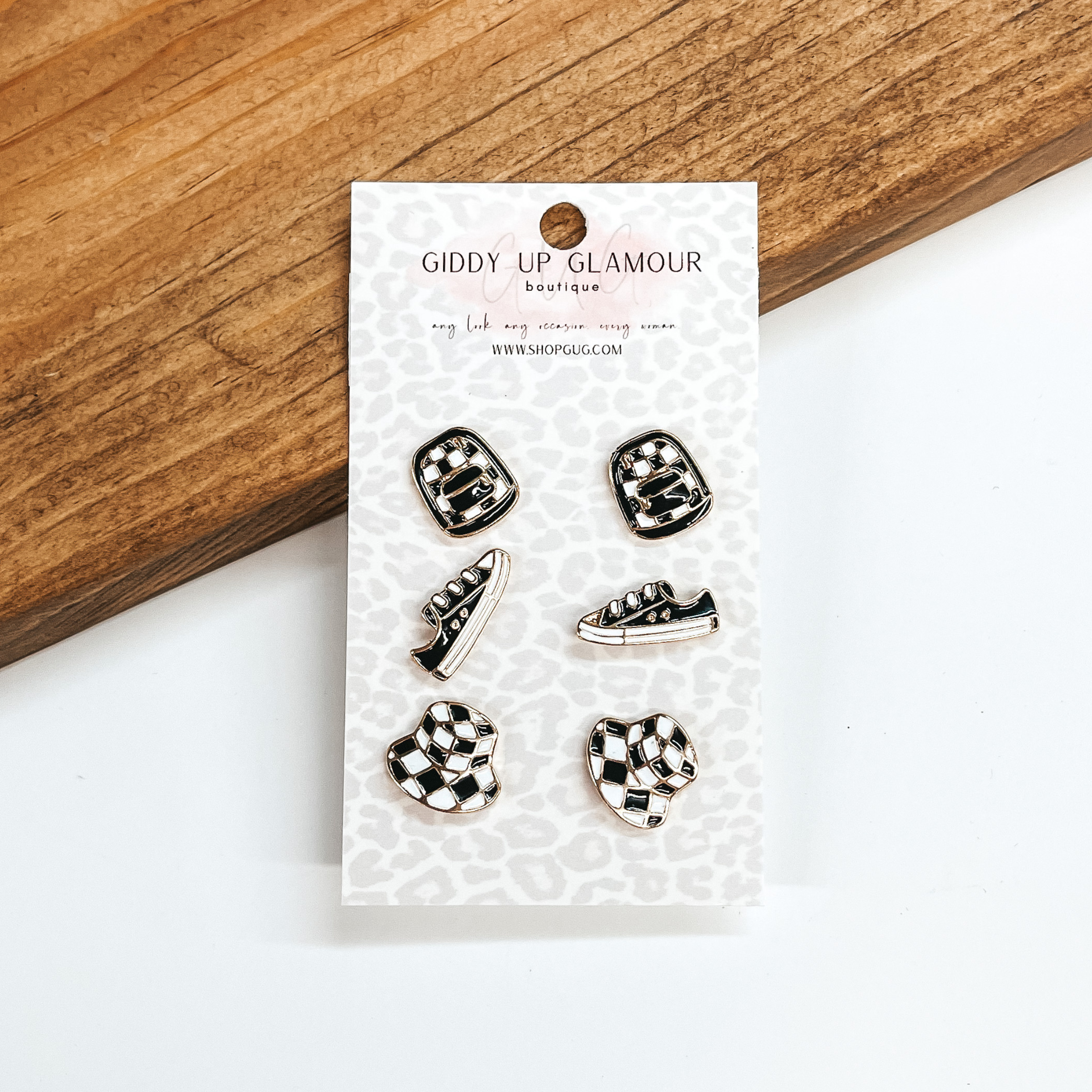 These are three set of stud earrings in black and  white checkered pattern. The bottom set are  bucket hats, the middle set are sneakers, and the  top set are backpacks. These earrings are taken on  a Giddy Up Glamour cardboard, a brown block and a  white background. 