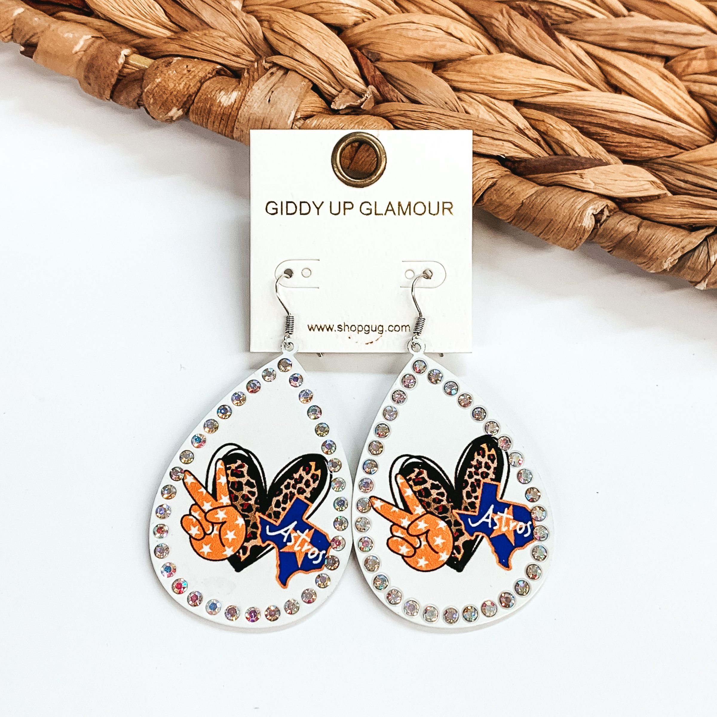 These are white teardrop earrings with AB crystals  all around. It has a leopard print heart, a blue Texas  with Astros across, and an orange star print peace  sign. These earrings are taken on  a white background and leaned up against a brown  woven slate.