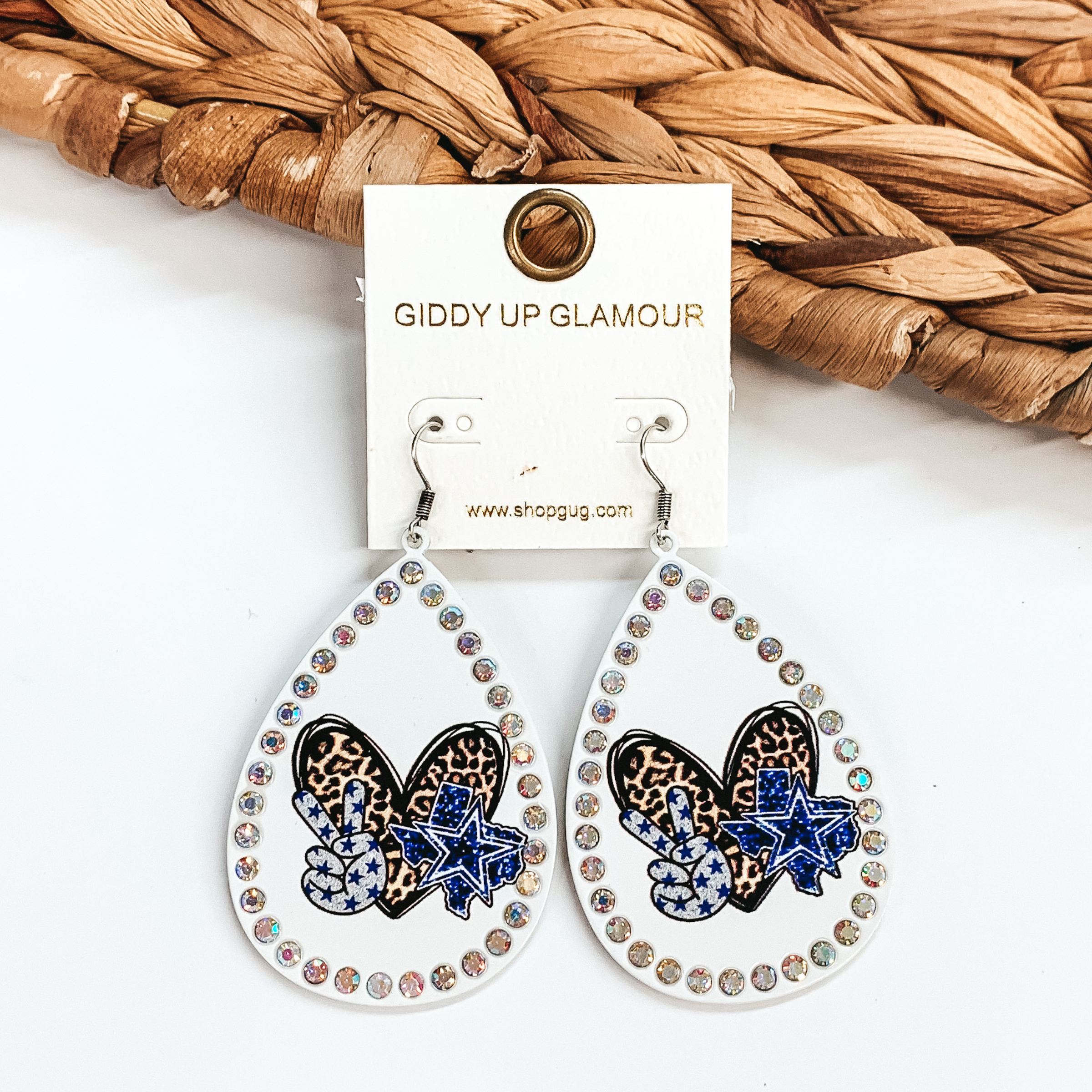 These are white teardrop earrings with AB crystals  all around. It has a leopard print heart, a blue Texas  with a blue heart in the middle, and a grey peace  sign with blue stars. These earrings are taken on  a white background and leaned up against a brown  woven slate.