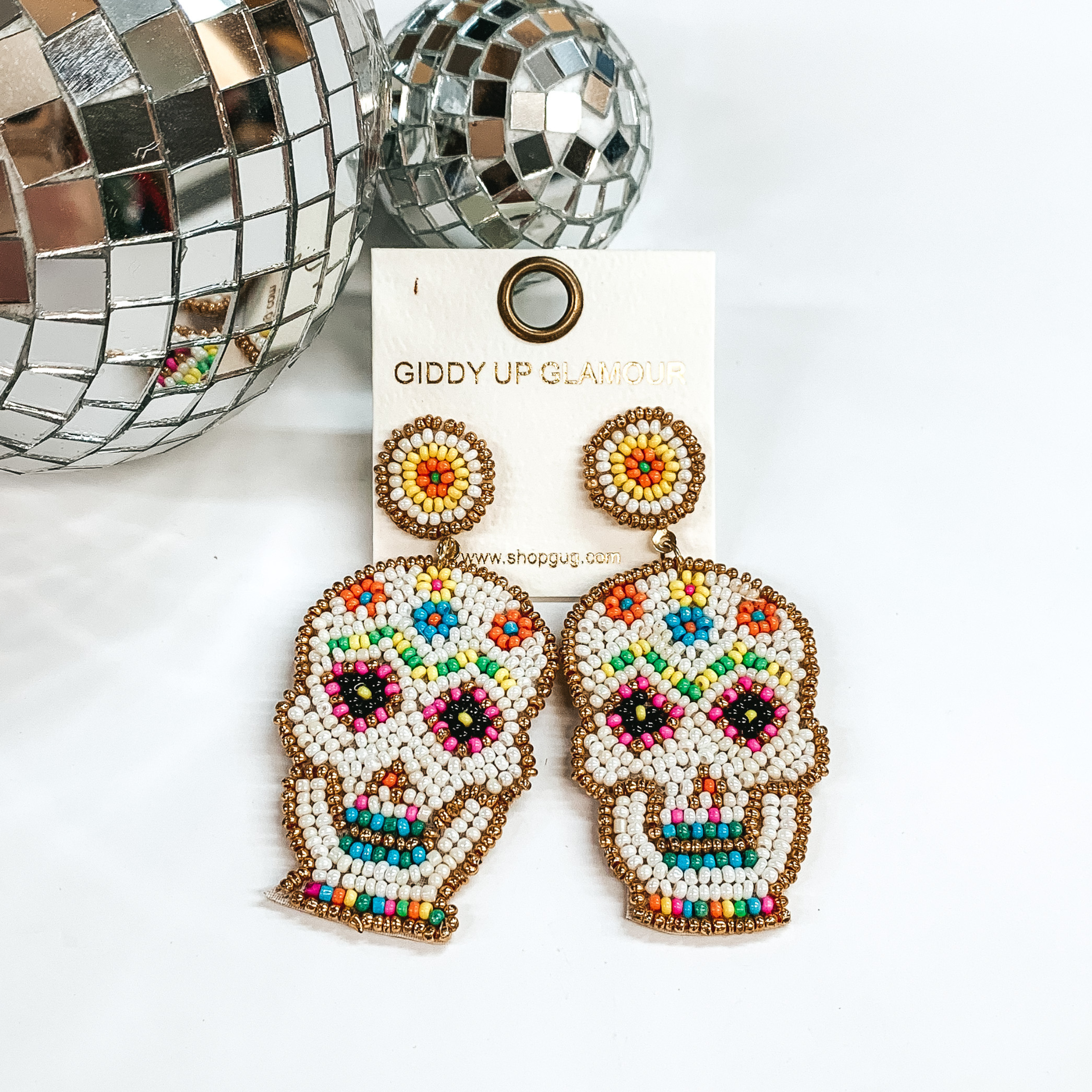 These are seedbeaded sugar skull post earrings in  white with multicolored beads as well. These earrings  are taken on a white background and two disco balls  in the back as decor.
