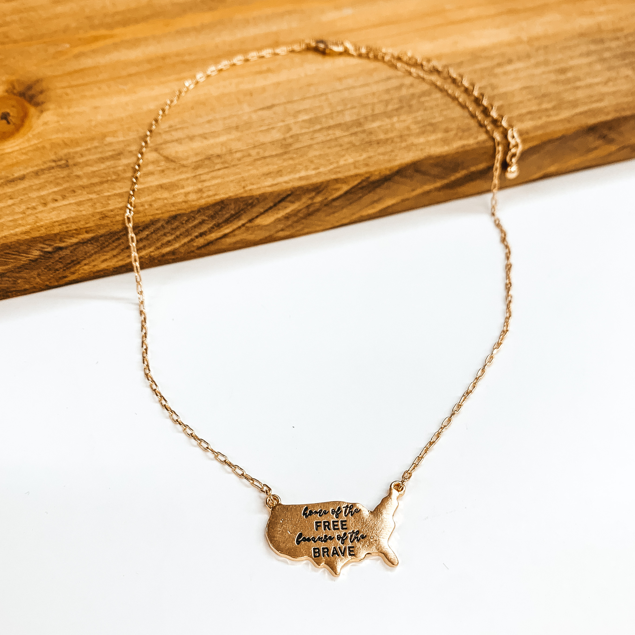 This a gold necklace with the USA country pendant,  the pendant says 'Home of the free because of the  brave'. This necklace is taken on a brown block  and a white background.
