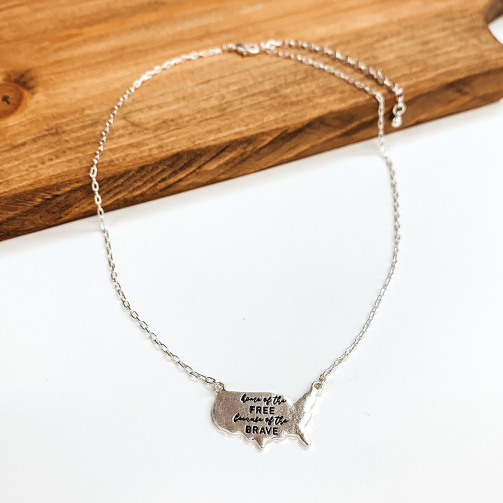 This a silver necklace with the USA country pendant,  the pendant says 'Home of the free because of the  brave'. This necklace is taken on a brown block  and a white background.