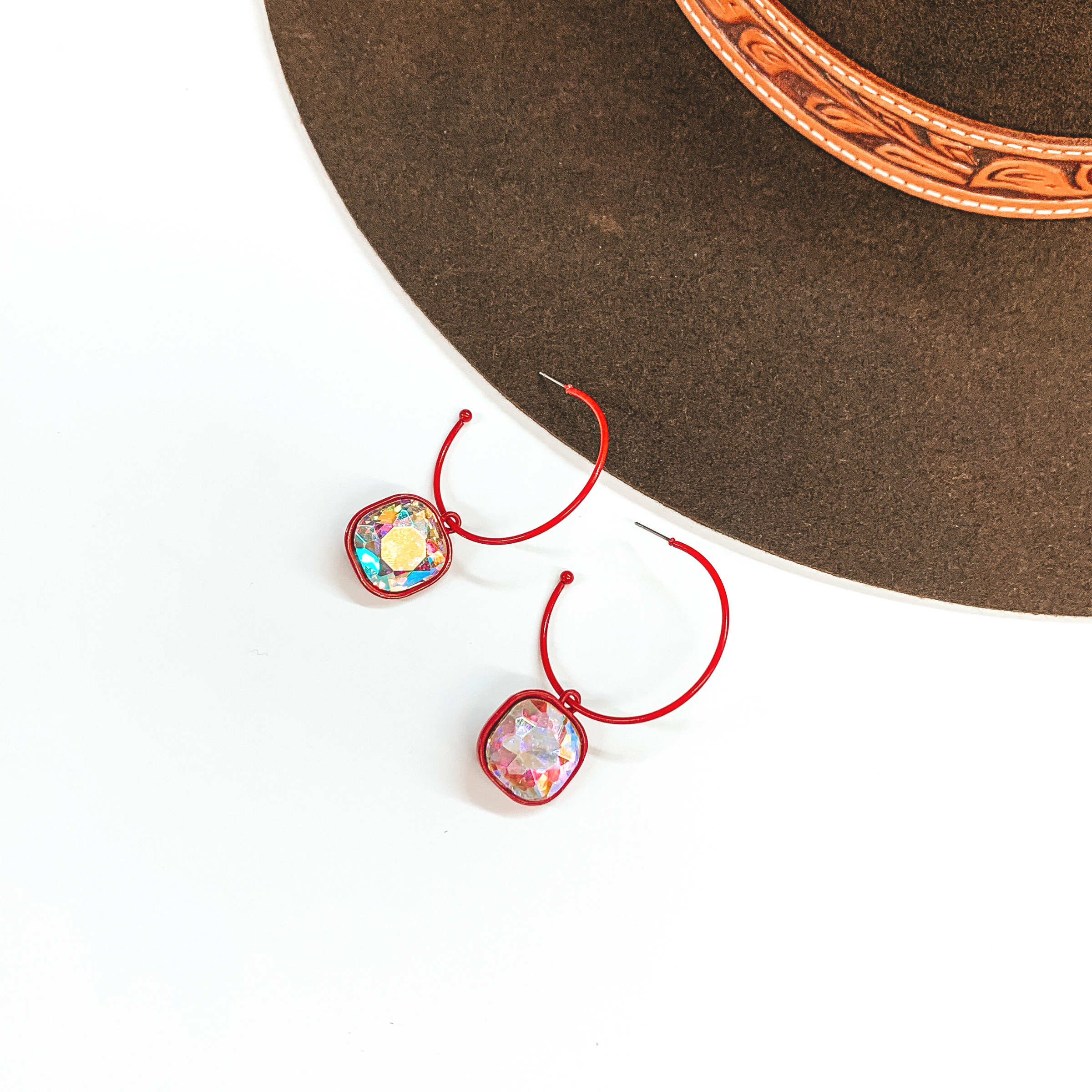 These are red open ended hoop earrings with  a cushion cut AB crystal hanging pendant. These  earrings are taken on a white background and a dark  brown hat in the back as decor.