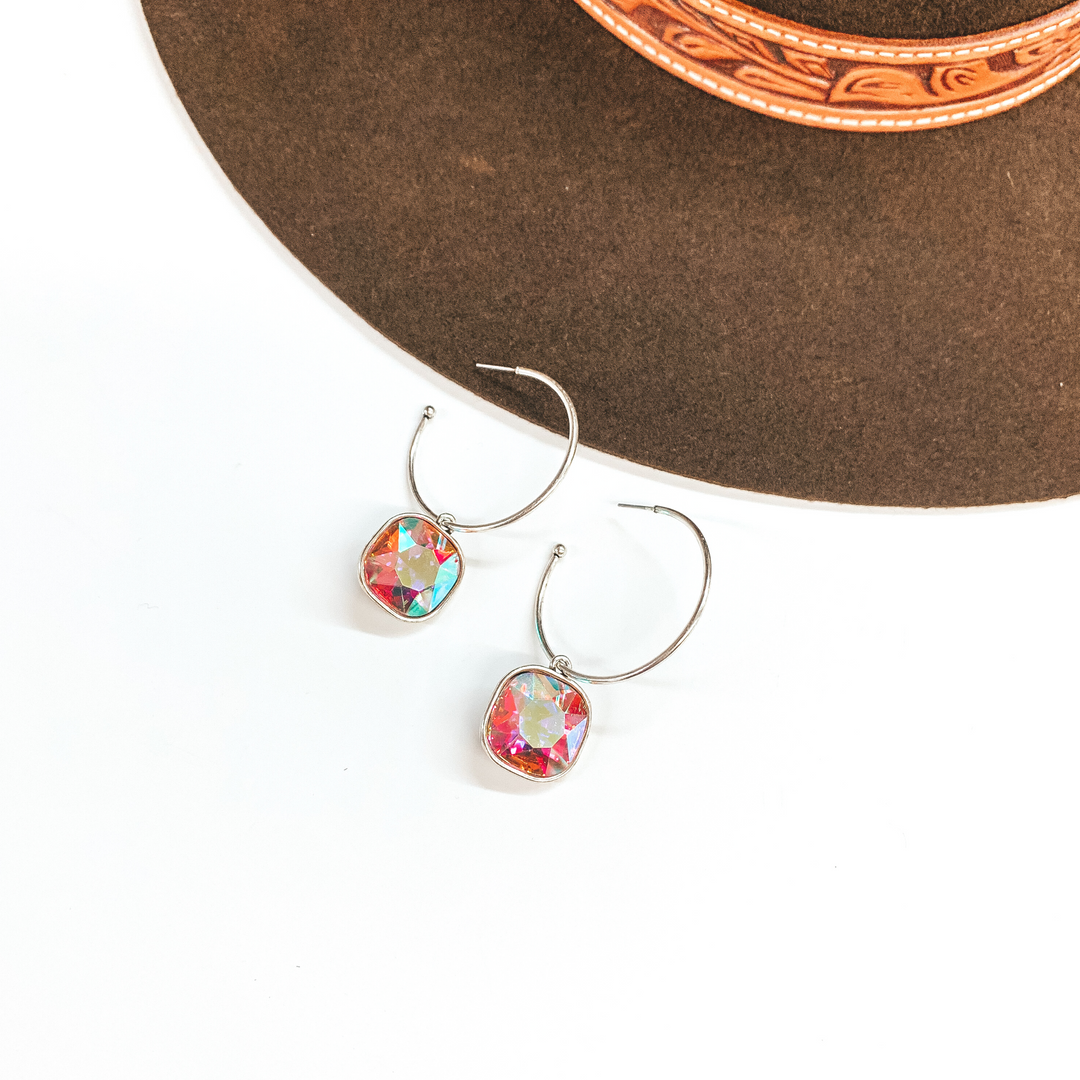 These are silver open ended hoop earrings with  a cushion cut AB crystal hanging pendant. These  earrings are taken on a white background and a dark  brown hat in the back as decor.