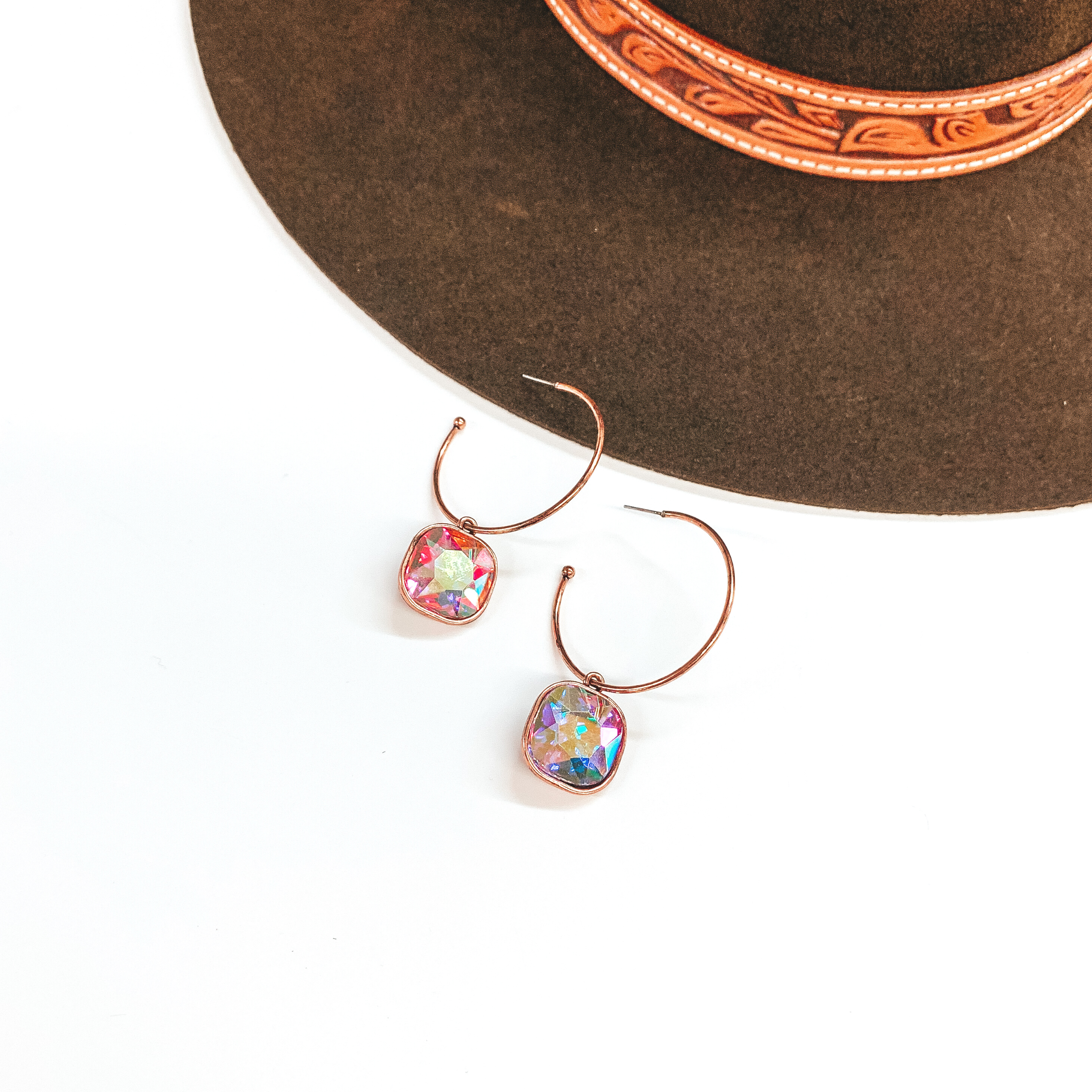 These are copper open ended hoop earrings with  a cushion cut AB crystal hanging pendant. These  earrings are taken on a white background and a dark  brown hat in the back as decor.