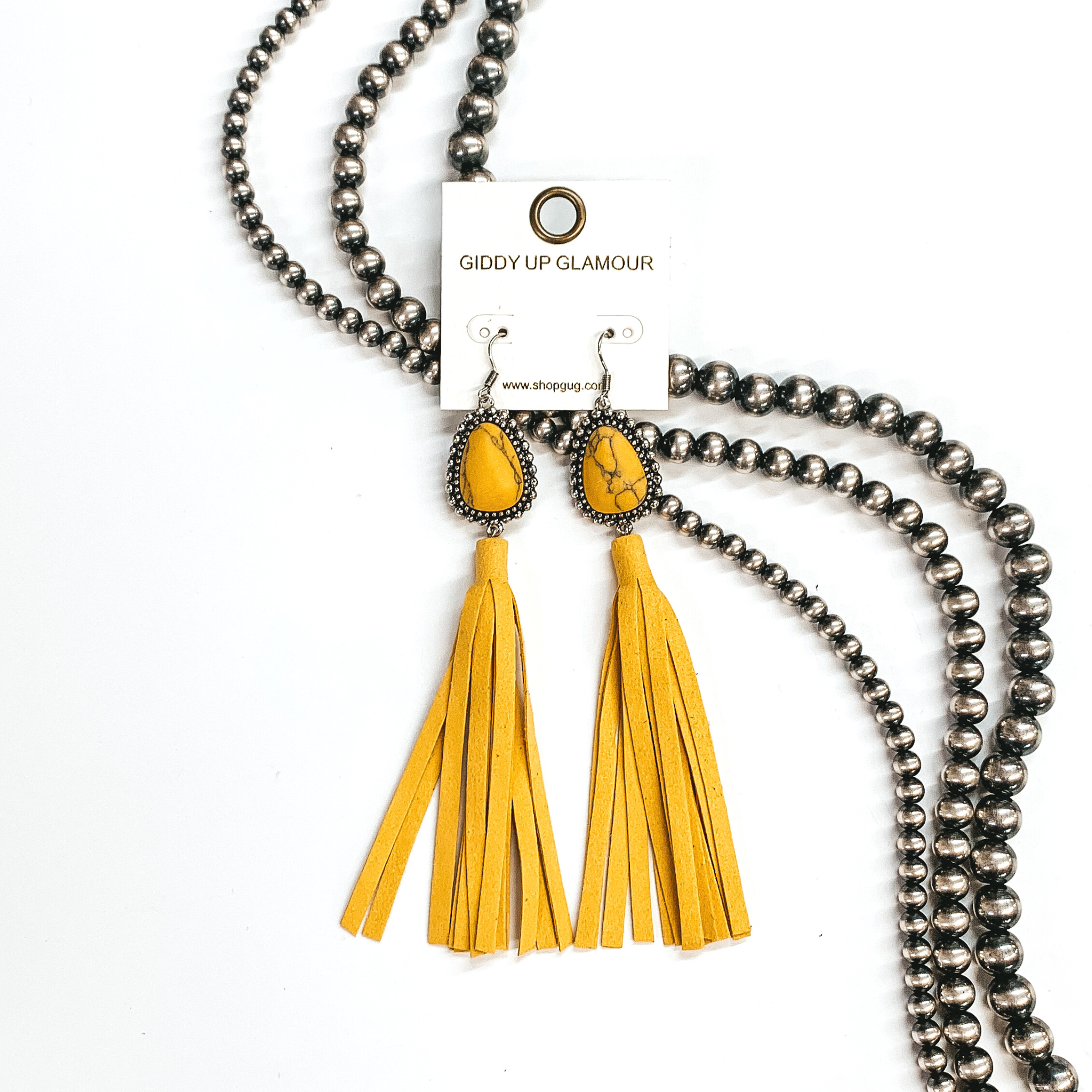 Yellow teardrop stone earrings in a silver setting with a faux leather  tassels in yellow. These earrings are taken on a white background and  Navajo pearls in the back as decor.