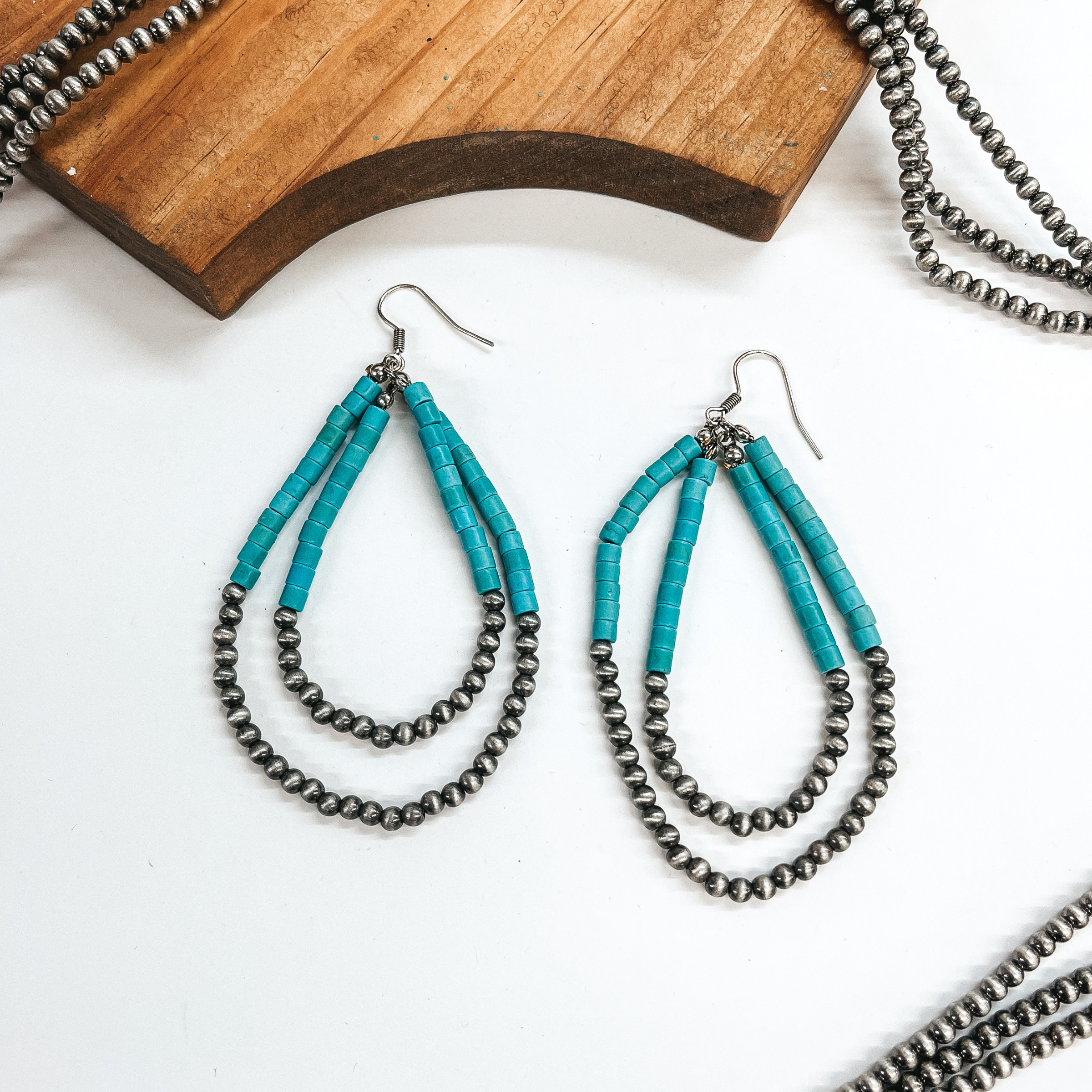 Double layered teardrop earrings, top half has turquoise beads and  bottom half with silver Navajo beads. These earrings are taken laying on  a white background with silver Navajo beads in the sides as decor and a  brown block.