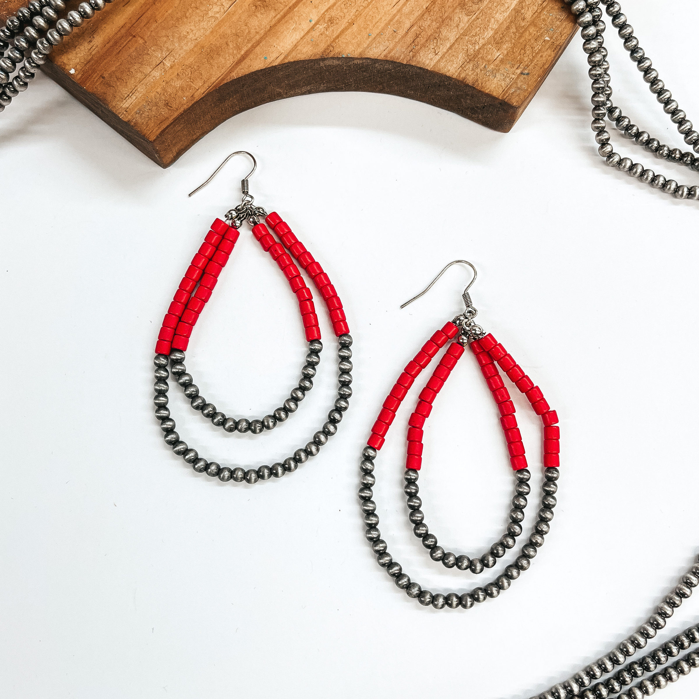 Double layered teardrop earrings, top half has red beads and  bottom half with silver Navajo beads. These earrings are taken laying on  a white background with silver Navajo beads in the sides as decor and a  brown block.