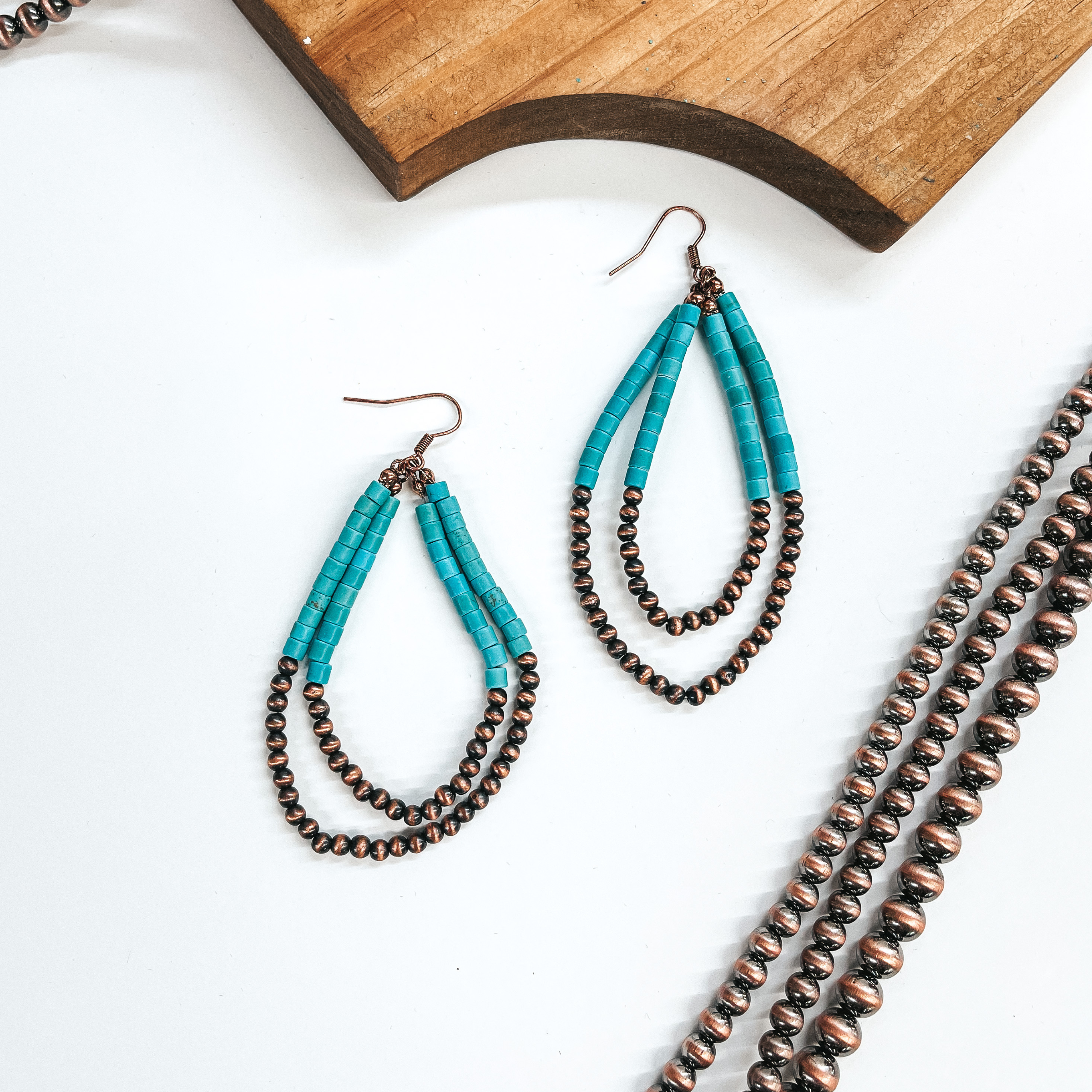 Double layered teardrop earrings, top half has turquoise beads and  bottom half with copper Navajo beads. These earrings are taken laying on  a white background with copper Navajo beads in the sides as decor and a  brown block.