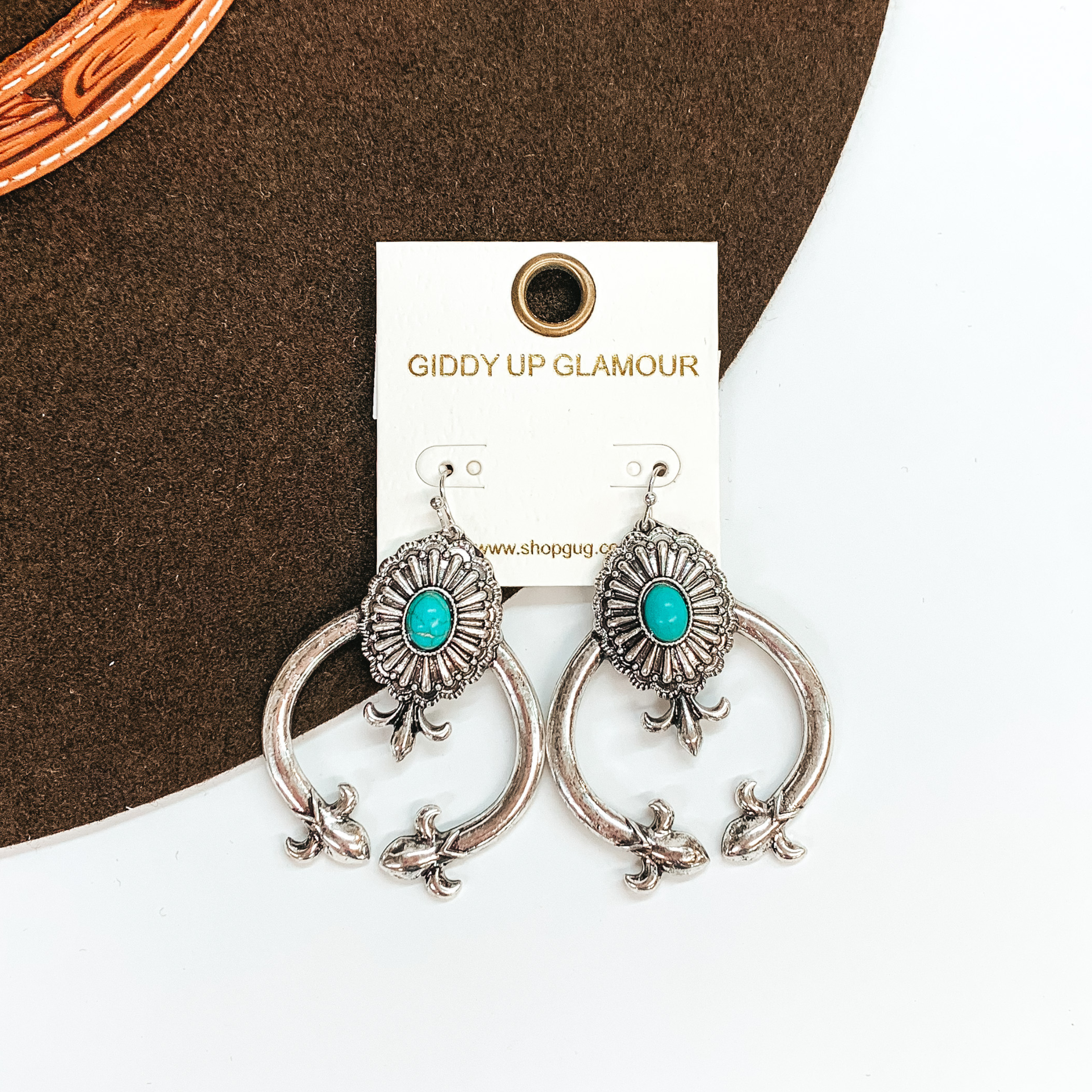 These are silver squash blossom earrings with a concho in the center and  a small oval faux stone in turquoise. The squash blossom have pointy ends and under the concho. These earrings are taken laying on a dark brown  brim hat and on a white background.