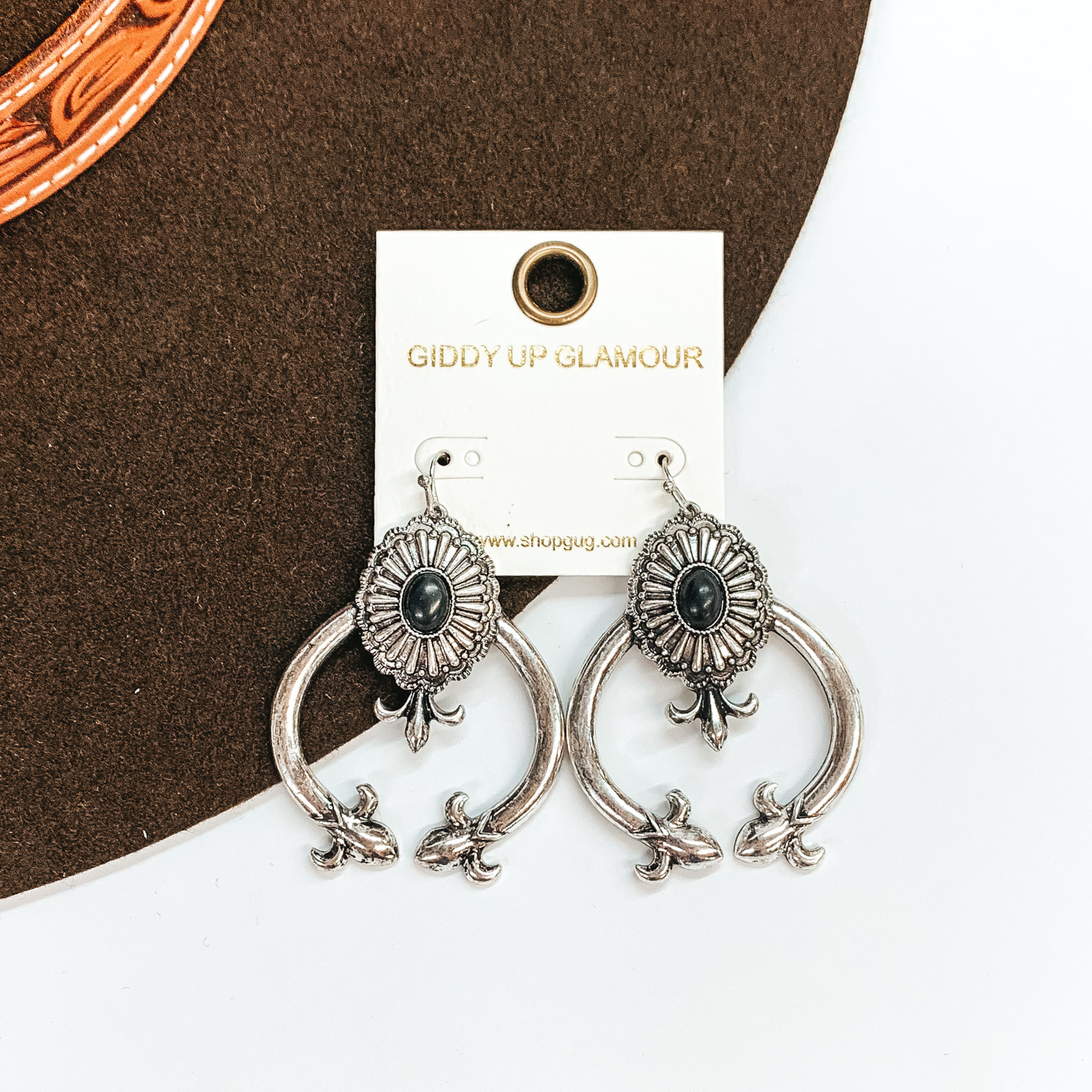 These are silver squash blossom earrings with a concho in the center and  a small oval faux stone in black. The squash blossom have pointy ends and under the concho. These earrings are taken laying on a dark brown  brim hat and on a white background.