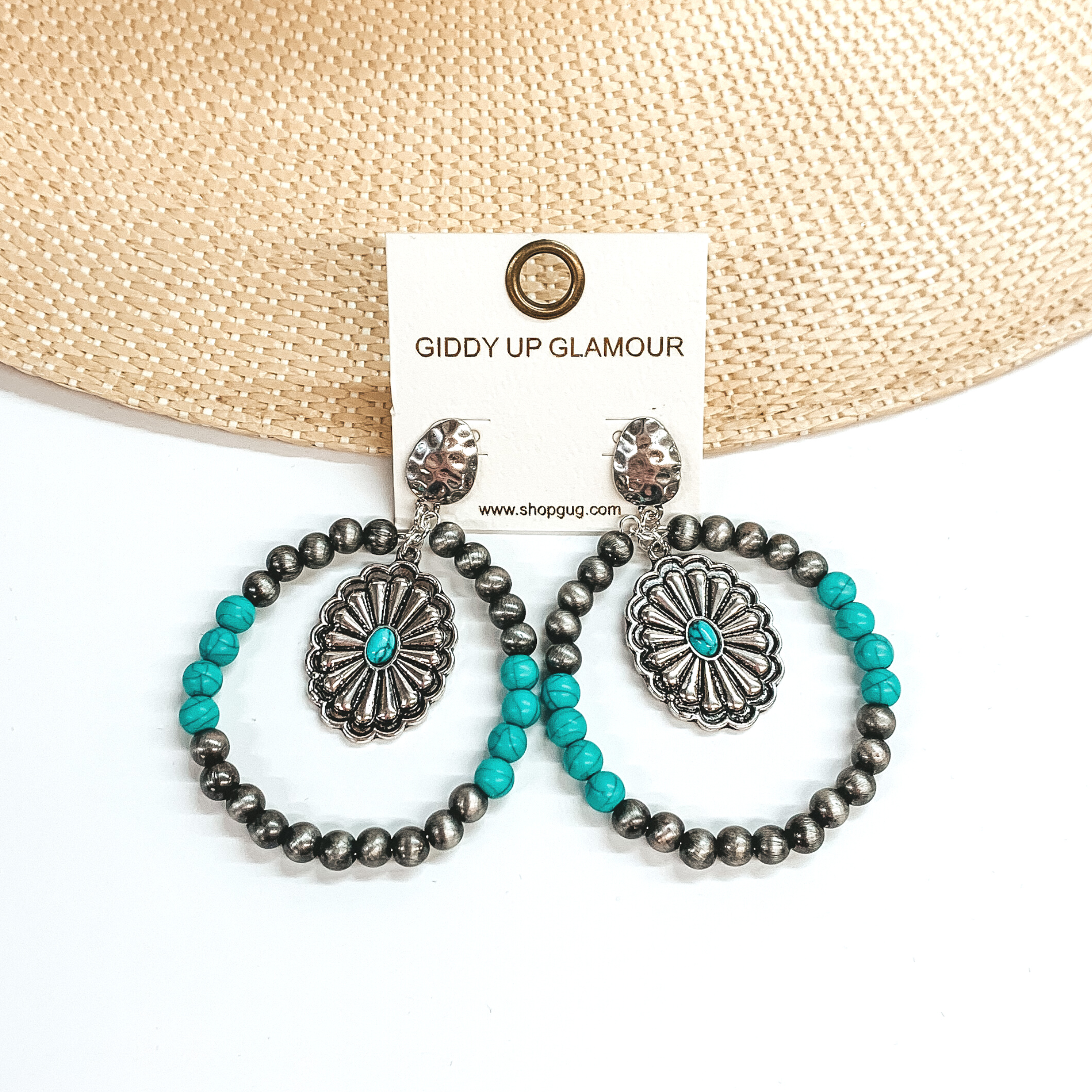 These are silver and turquoise beaded circle drop earrings with a  silver hammered post back. The circle drop has Navajo pearls in silver  and four turquoise beads in each side. There is a silver concho pendant  in the middle with a small oval turquoise stone.  These earrings are taken leaning on a light brown  straw hat brim and a white background.