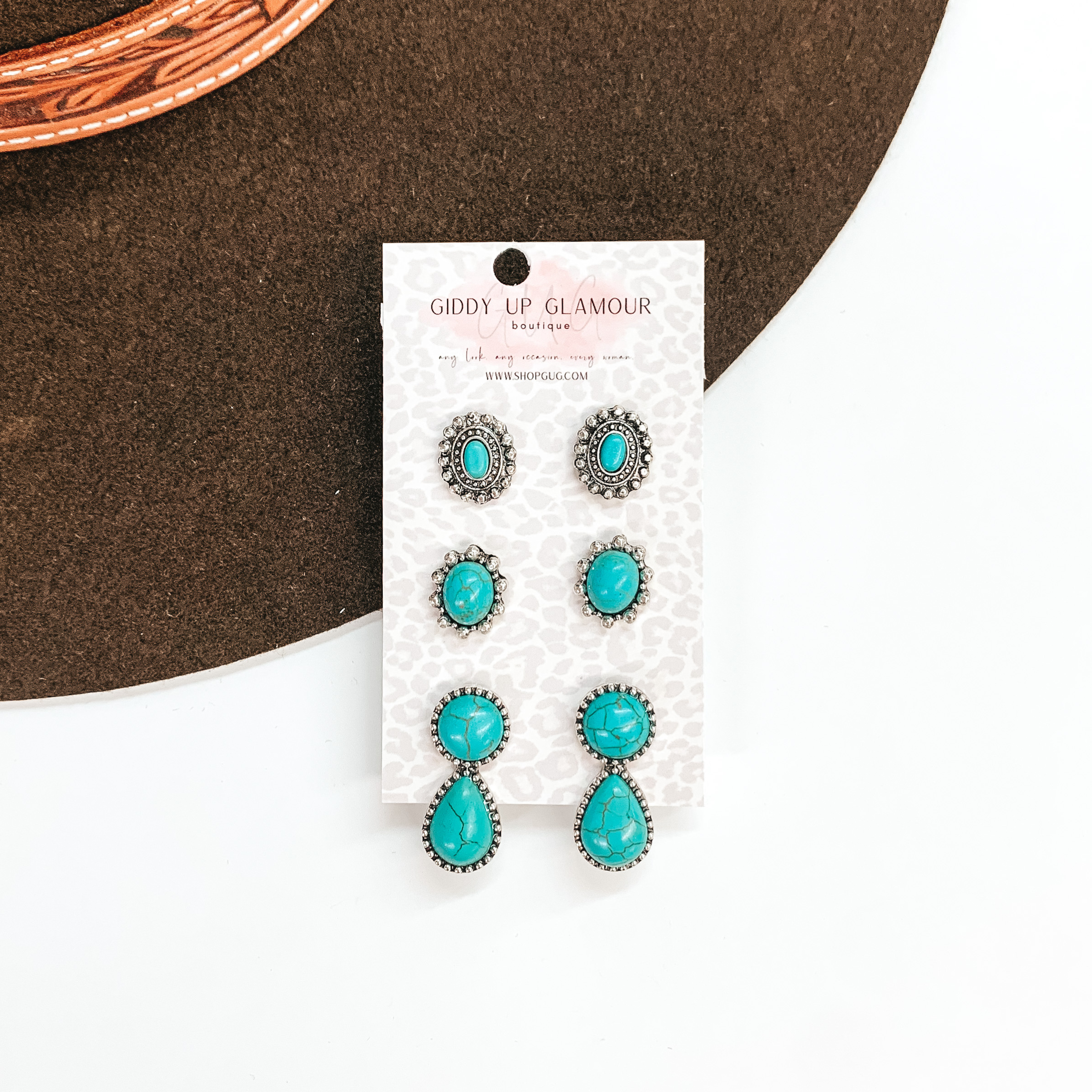 Set of three turquoise and silver earrings in different sizes. The top  pair is a silver concho with a small stone in the center. The middle pair  is a larger oval stone with silver pointy edges. The bottom pair has a  circle stone connected to a teardrop shaped stone in a silver setting.  These earrings are placed on a white  and grey leopard card holder, the earrings are taken on a dark brown hat  brim and a white background.