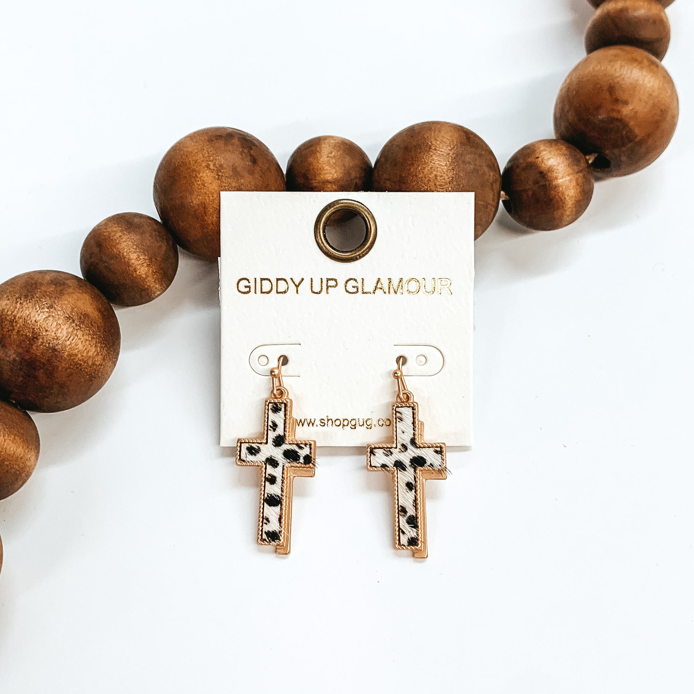 Gold cross earrings with a faux hide inlay in black and white dotted print. They have a gold shadow as well. These earrings are taken leaning up  against dark brown beads and white background.