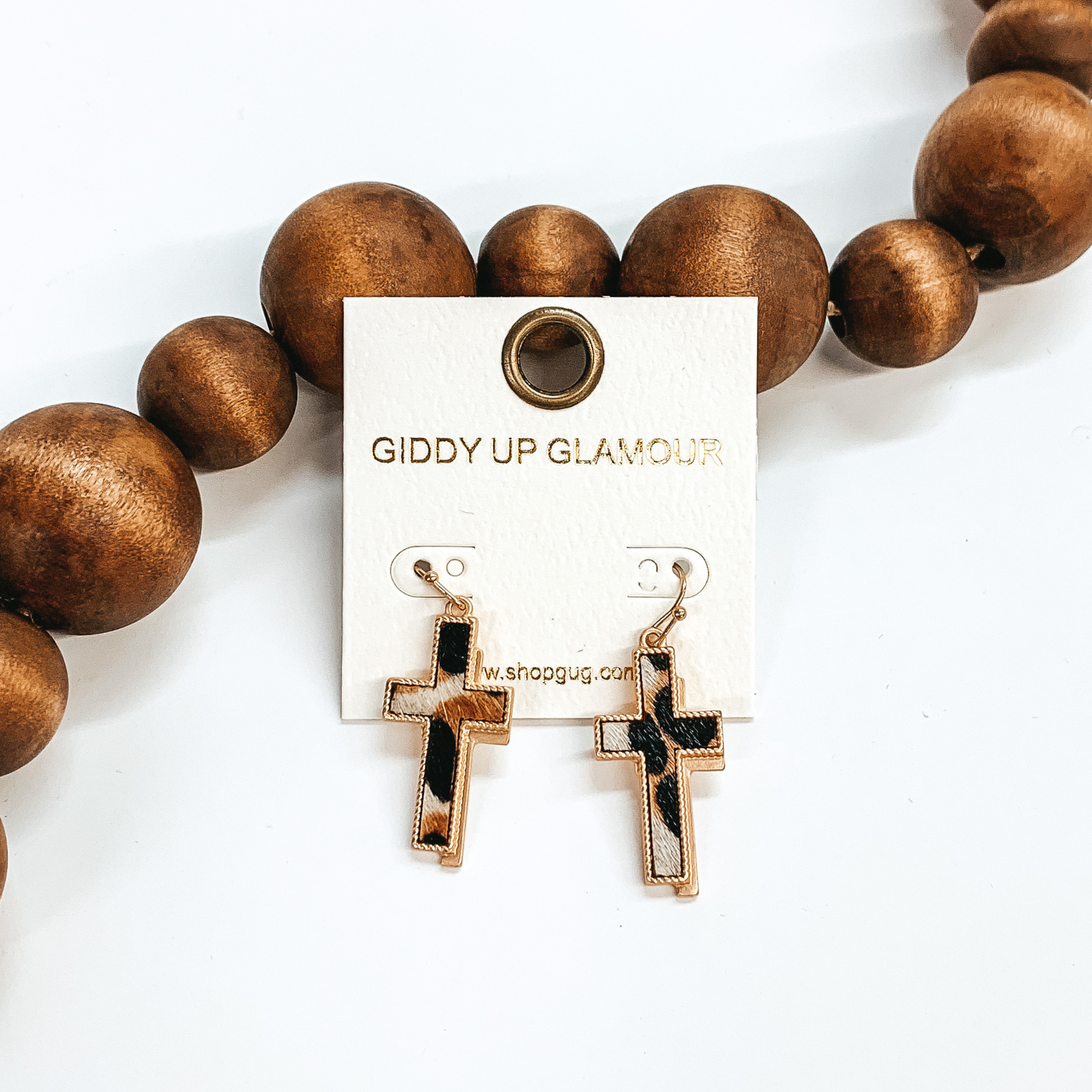 Gold cross earrings with a faux hide inlay in black, white, and brown print. They have a gold shadow as well. These earrings are taken leaning up  against dark brown beads and white background.