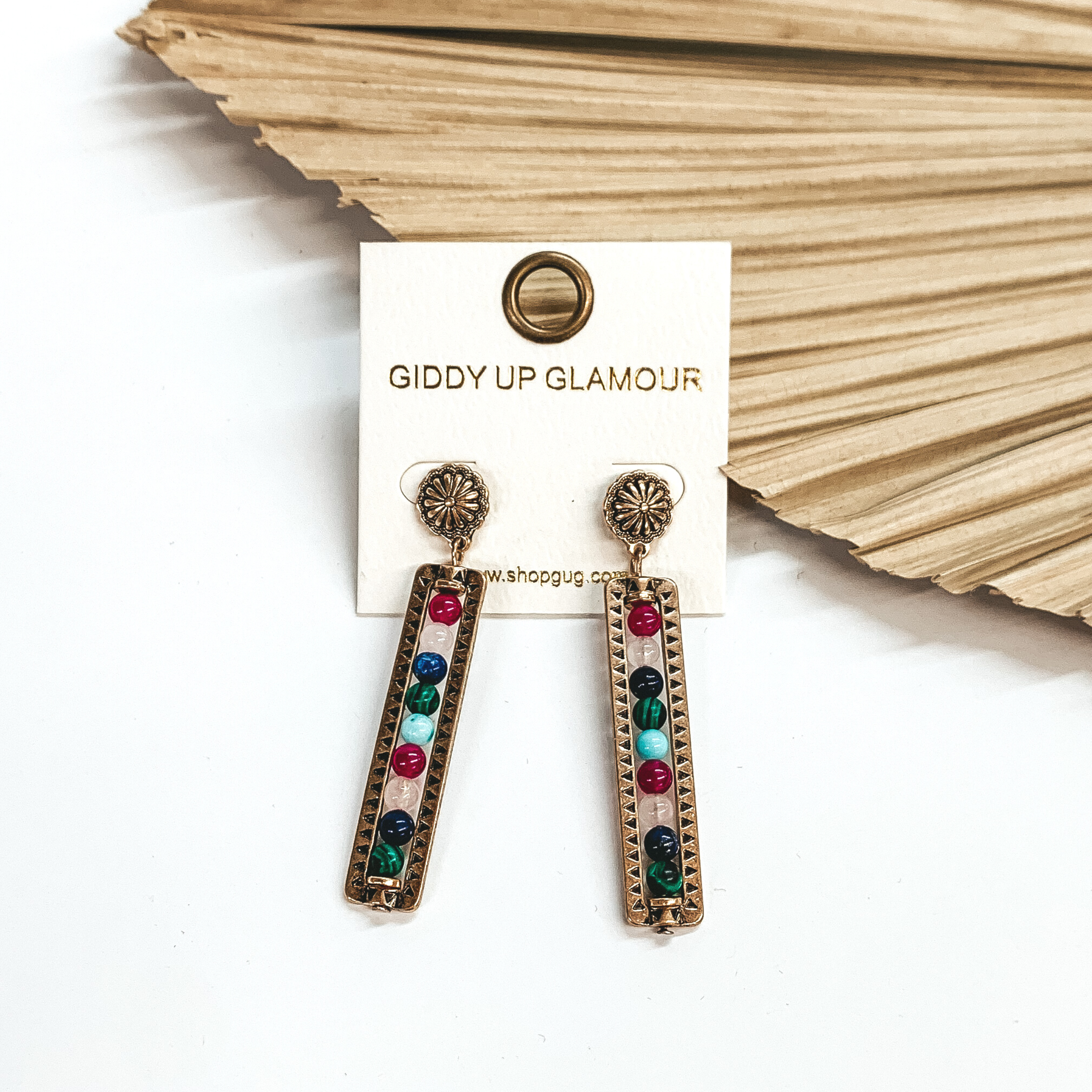 Gold small concho post earrings with a rectangle bar drop with  stone beads in multicolor and black detailing. Stone beads come in dark green,  navy blue, rose quarts, burgundy, and turquoise.  These earrings are taken leaning up  against a dried up palm leaf and a white background.