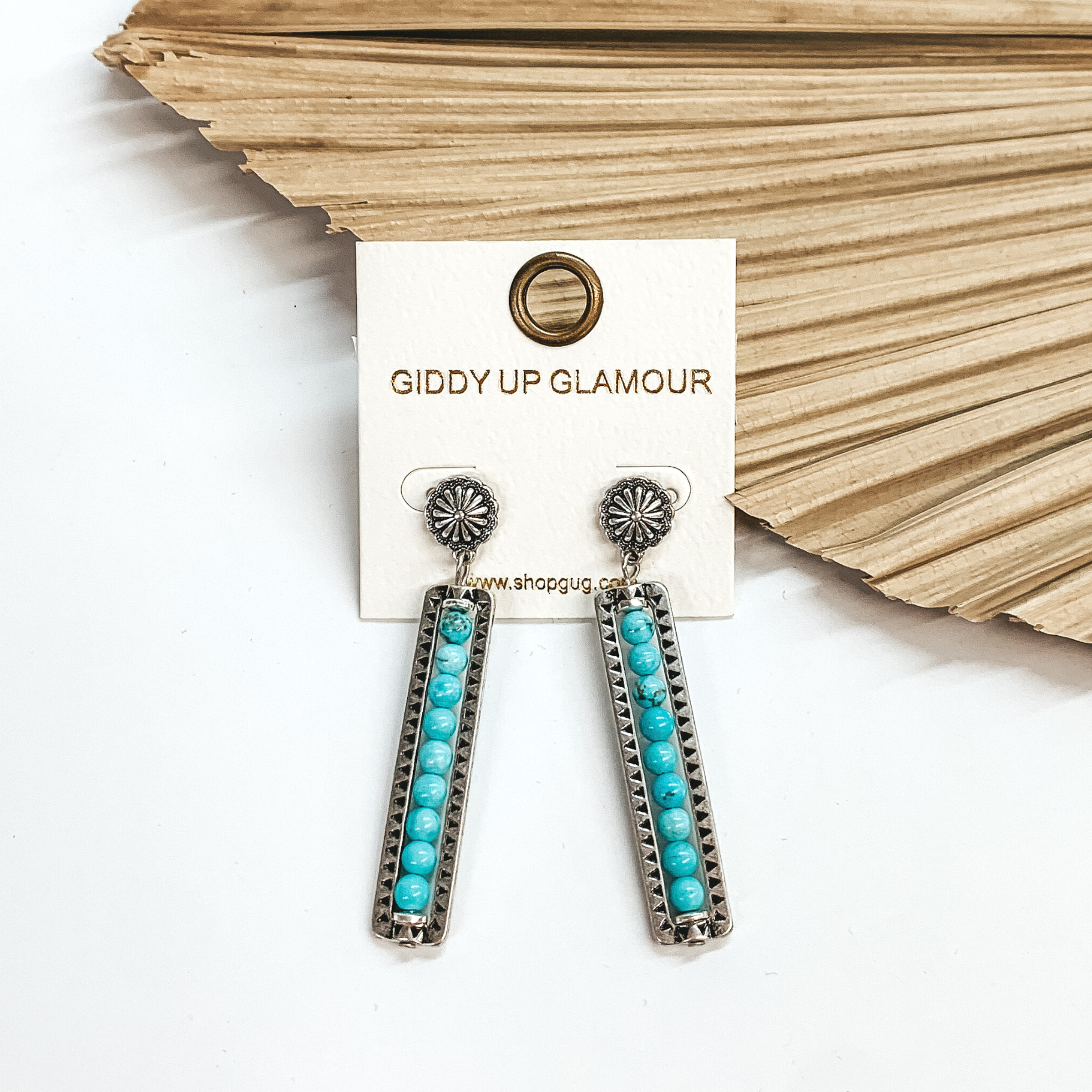 Silver small concho post earrings with a rectangle bar drop with  stone beads in turquoise and black detailing.  These earrings are taken leaning up  against a dried up palm leaf and a white background.
