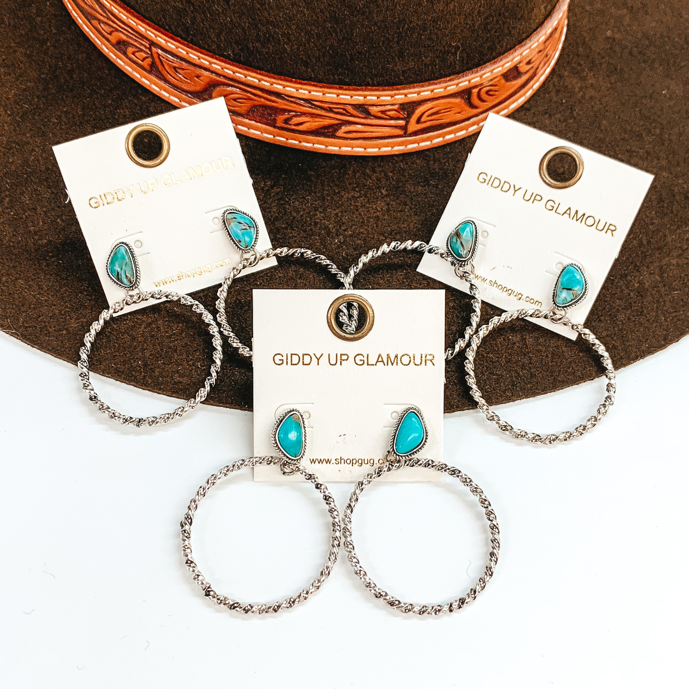 These are three pairs of silver circle drop earrings with an asymmetrical  turquoise stone post backing. The circle drop has a twisted  rope texture in silver. These earrings are taken laying on  a dark brown hat brim  and on a white background.