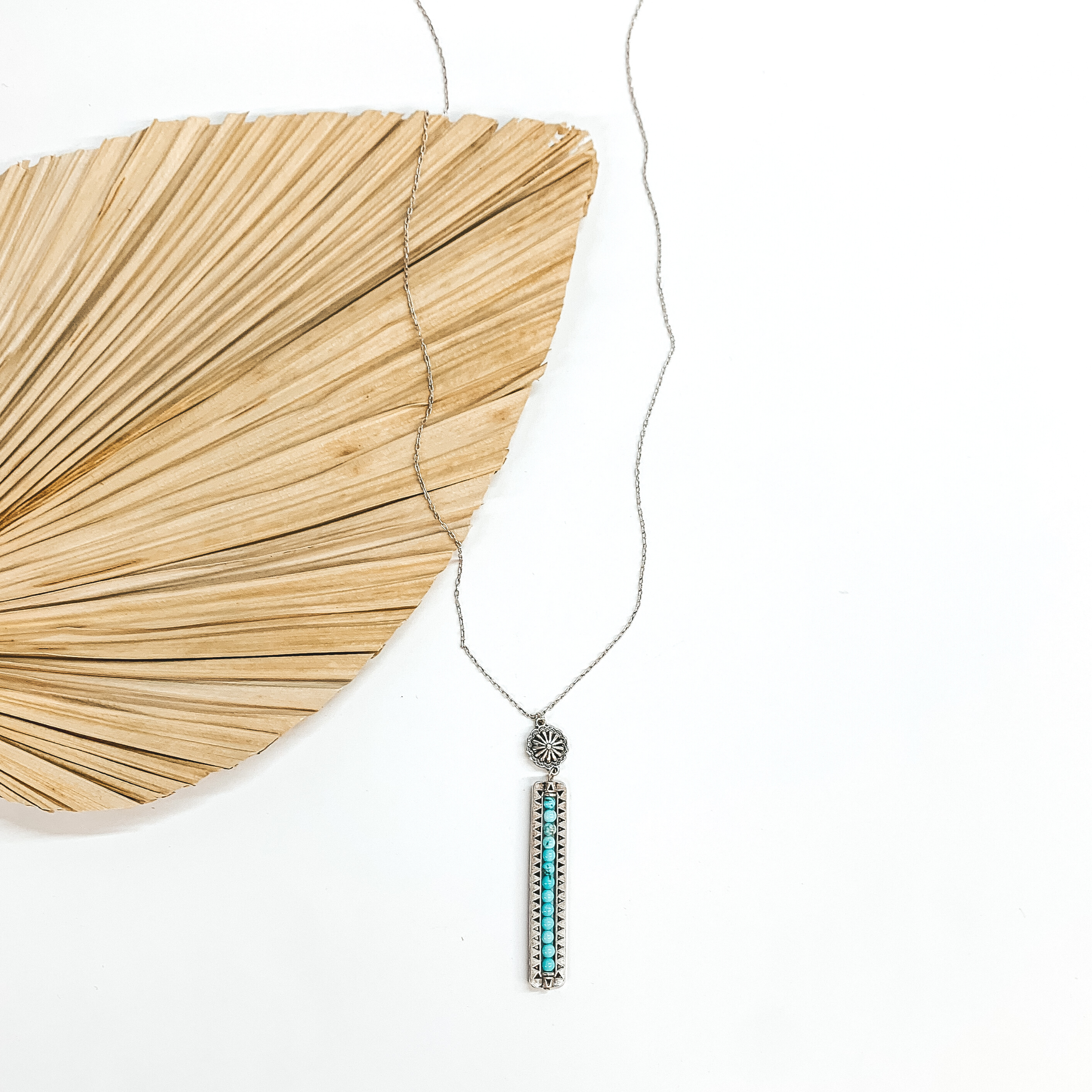 Long silver necklace with a concho and rectangle bar pendant. The  bar pendant has stone beads in turquoise and it has black details around.  This necklace is taken on a dried up palm leaf and a white background.
