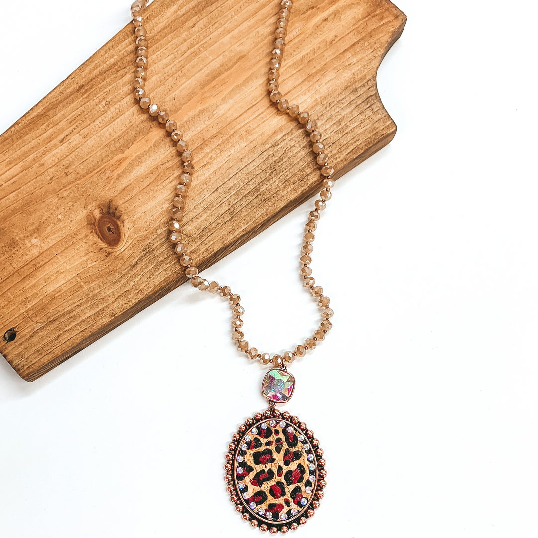 Long crystal beaded necklace in light brown with a leopard print pendant  and AB cushion cut crystal connector. The pendant is in a copper setting,  the leopard print is on a cork material and has small AB crystals all  around. This necklace is taken laying on a brown necklace board and white  background.