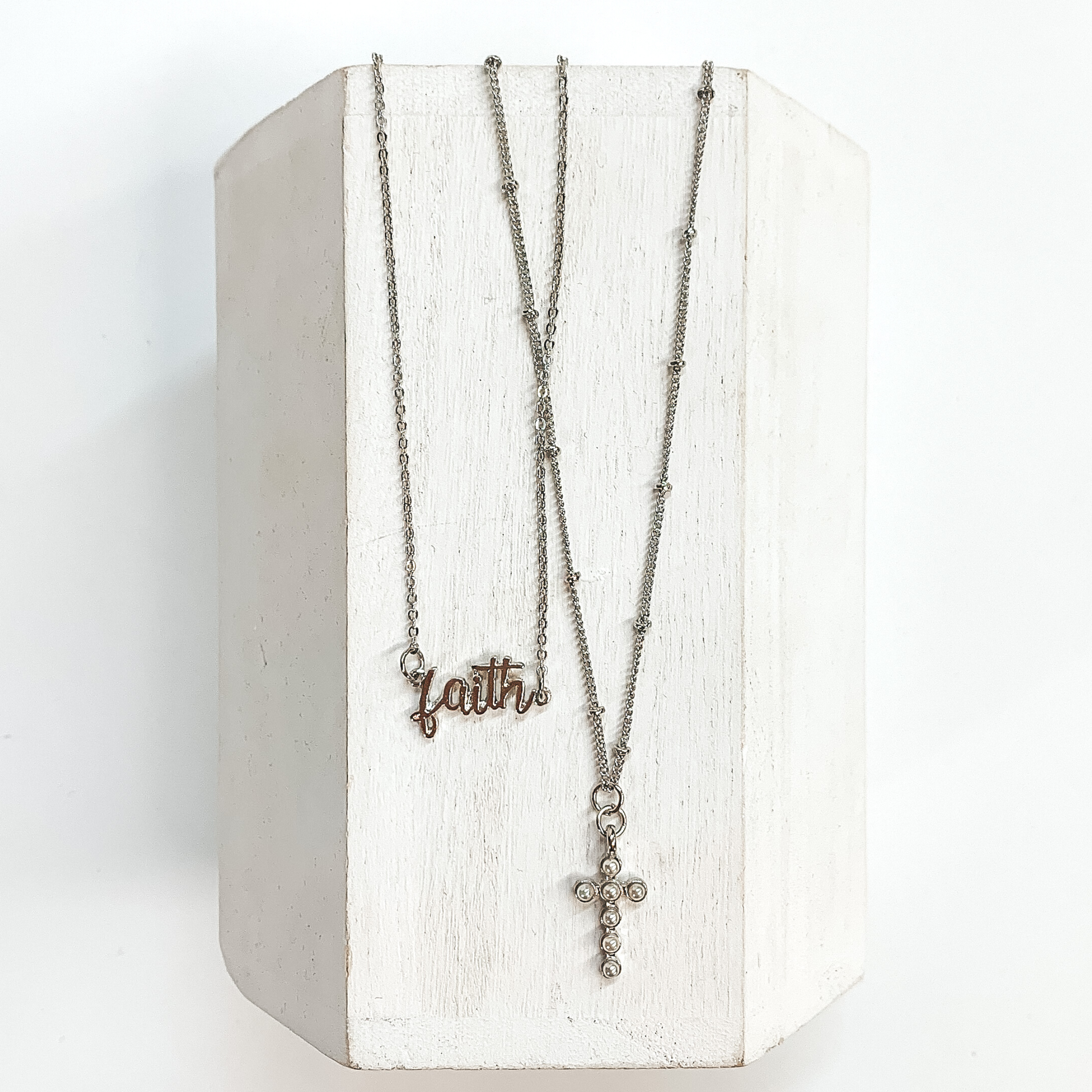Double layered silver necklace with a 'faith' pendant and cross pendant.  The longer necklace has silver spacers has a silver cross with pearls. The  shorter strand has 'faith' in the center. These necklaces are taken on a  white block and on a white background.