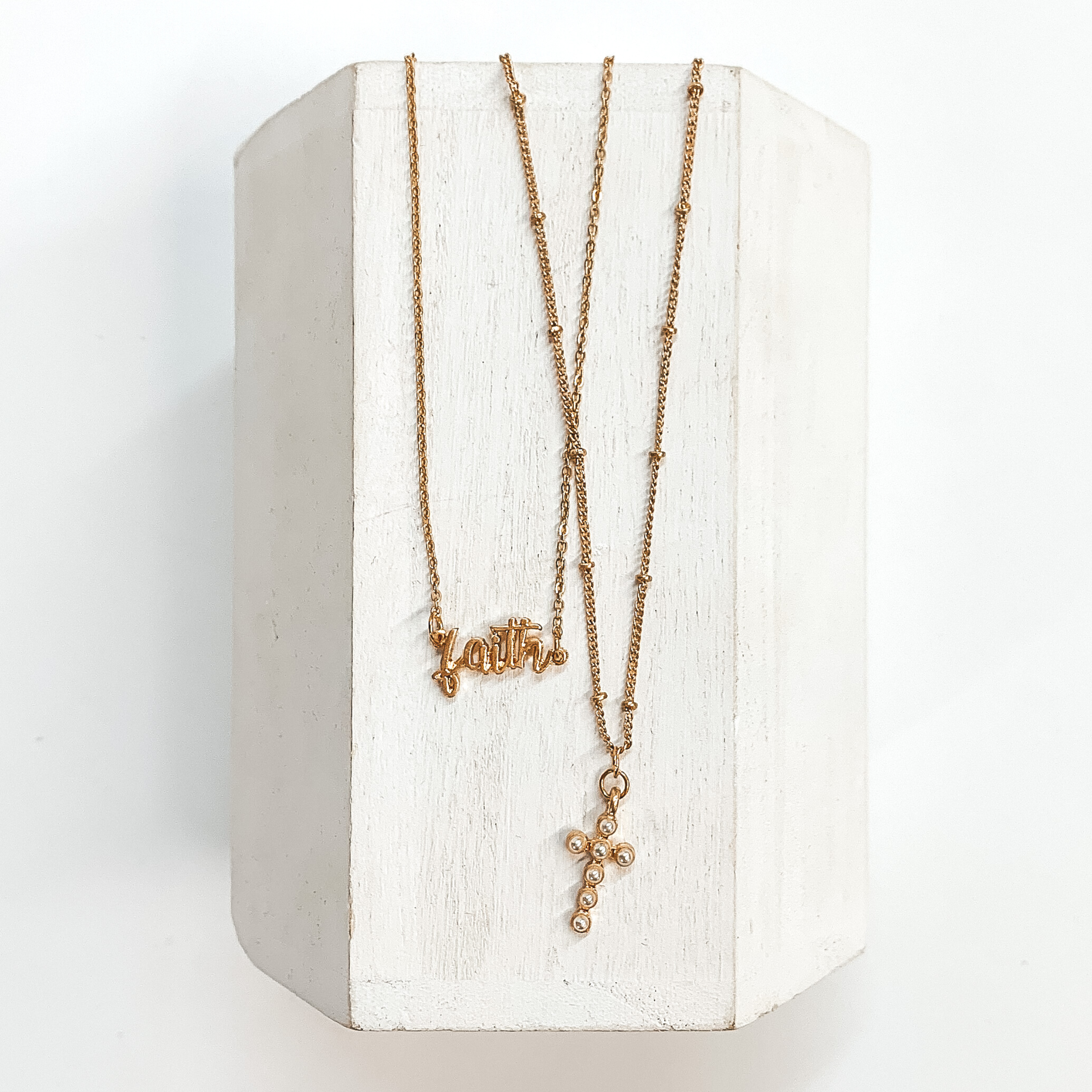 Double layered gold necklace with a 'faith' pendant and cross pendant.  The longer necklace has gold spacers has a gold cross with pearls. The  shorter strand has 'faith' in the center. These necklaces are taken on a  white block and on a white background.