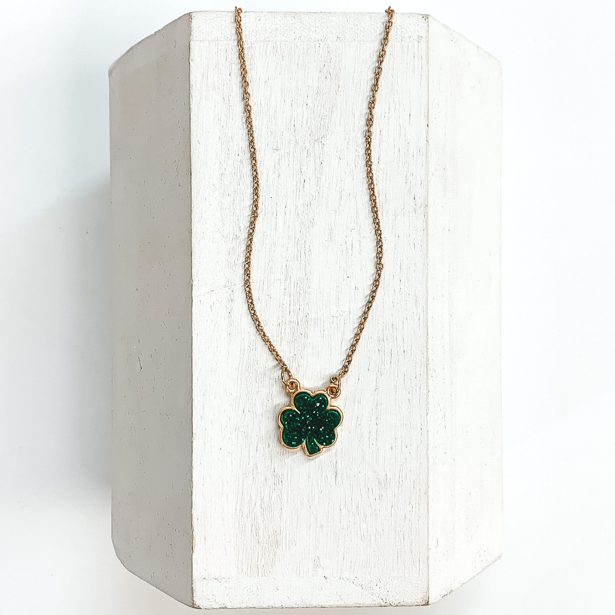 Gold chained necklace with green druzy four leaf clover pendant. This necklace is pictured laying on a white block on a white background.