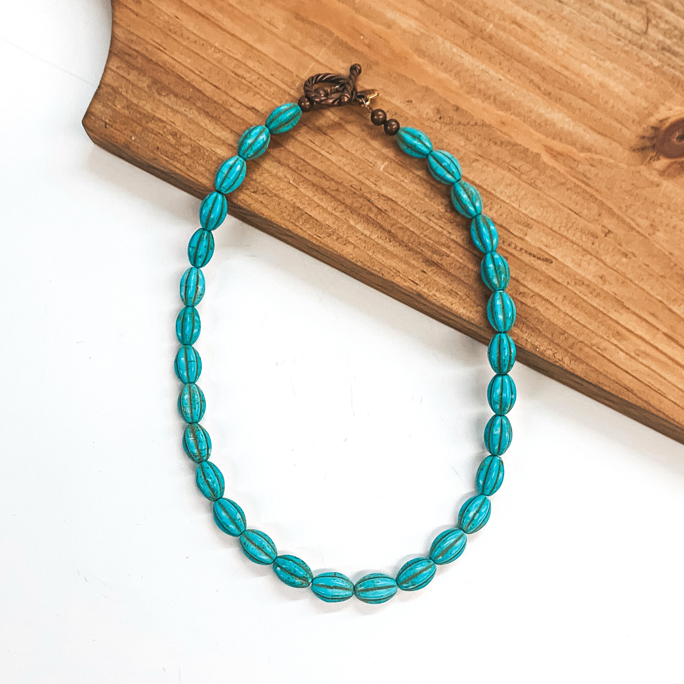 Turquoise beaded necklace with a copper toggle clasp.  This necklace is pictured partially laying on a brown necklace board and  on a white background.