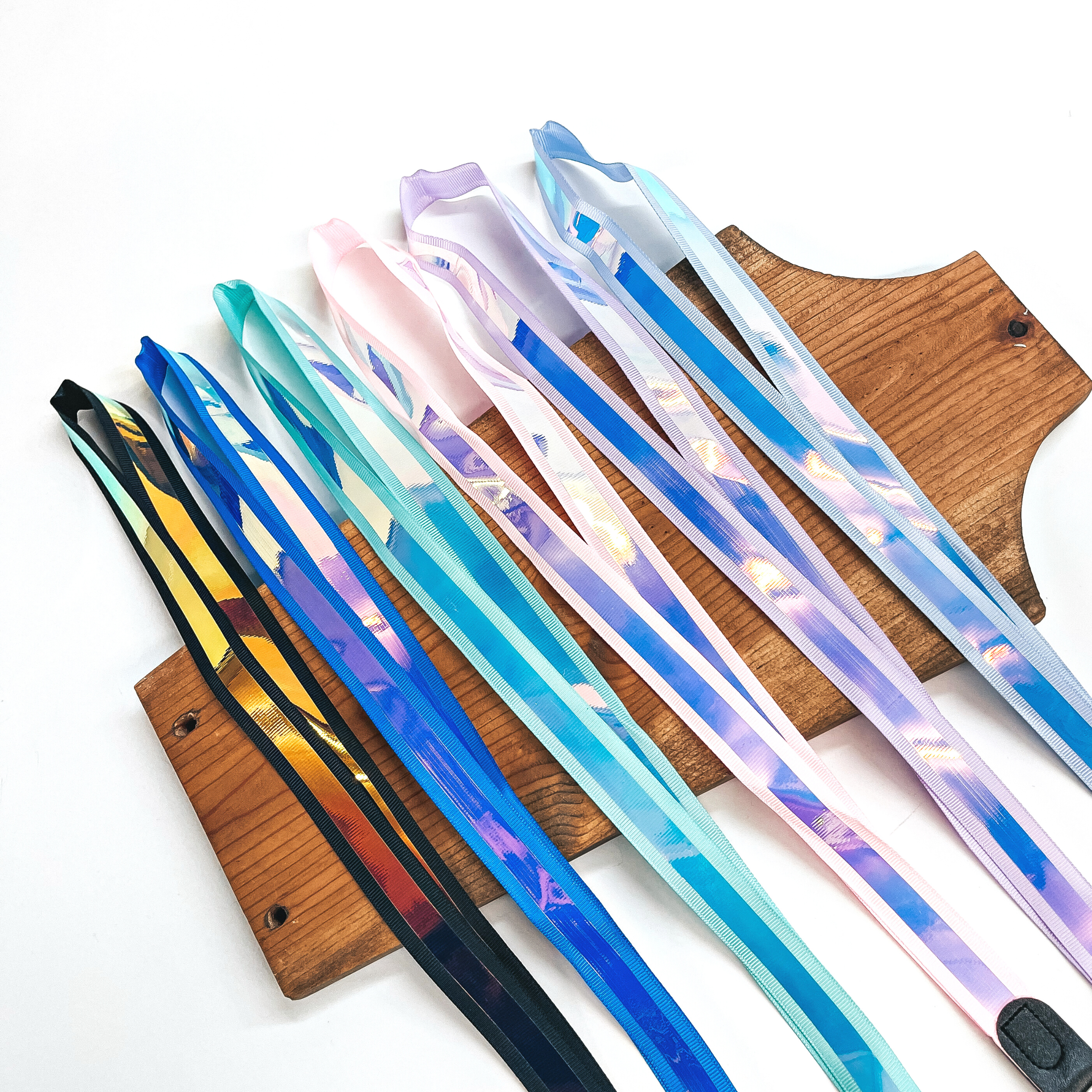There are six colored ribbon lanyards with a holographic ribbon in the  middle. From left to right: black, blue,  mint, pink, purple, and periwinkle. These lanyards are laying flat on a  brown necklace holder and on a white background.