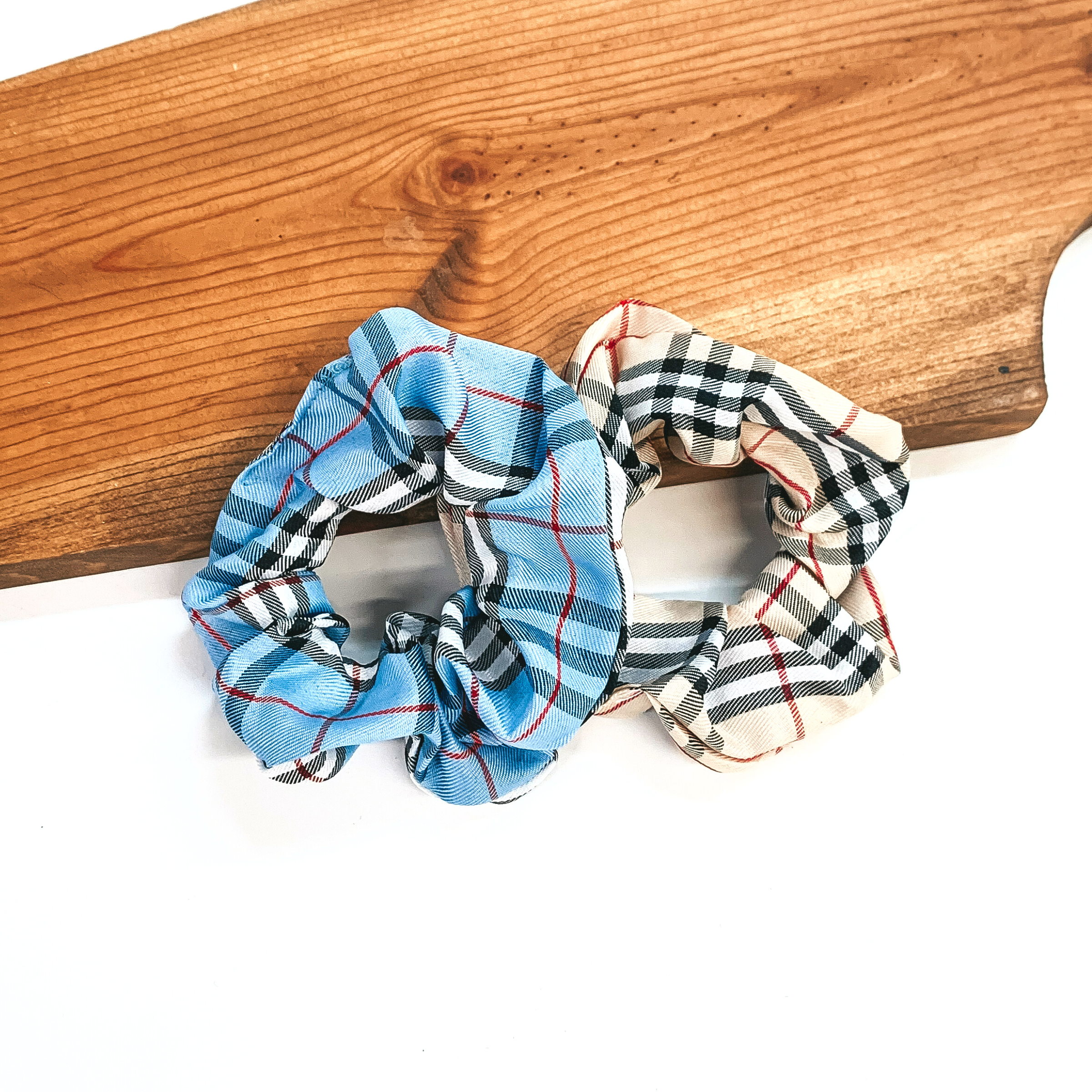 These are two plaid print scrunchies, the left one is light blue and right  one is in beige. Both of these scrunchies are taken leaning on a brown  necklace board and on a white background.