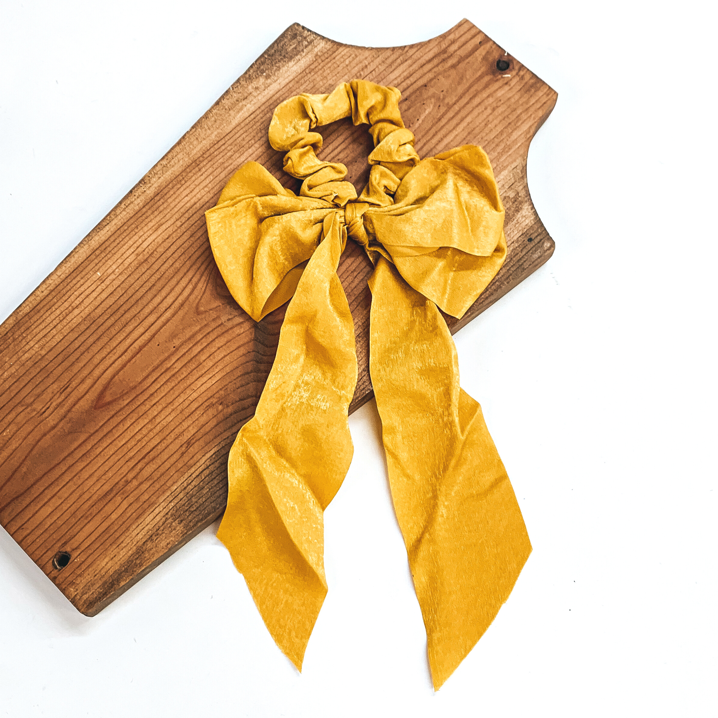 This is a mustard yellow satin scrunchie with a long bow, this scrunchie is taken on  a brown necklace board and on a white background.