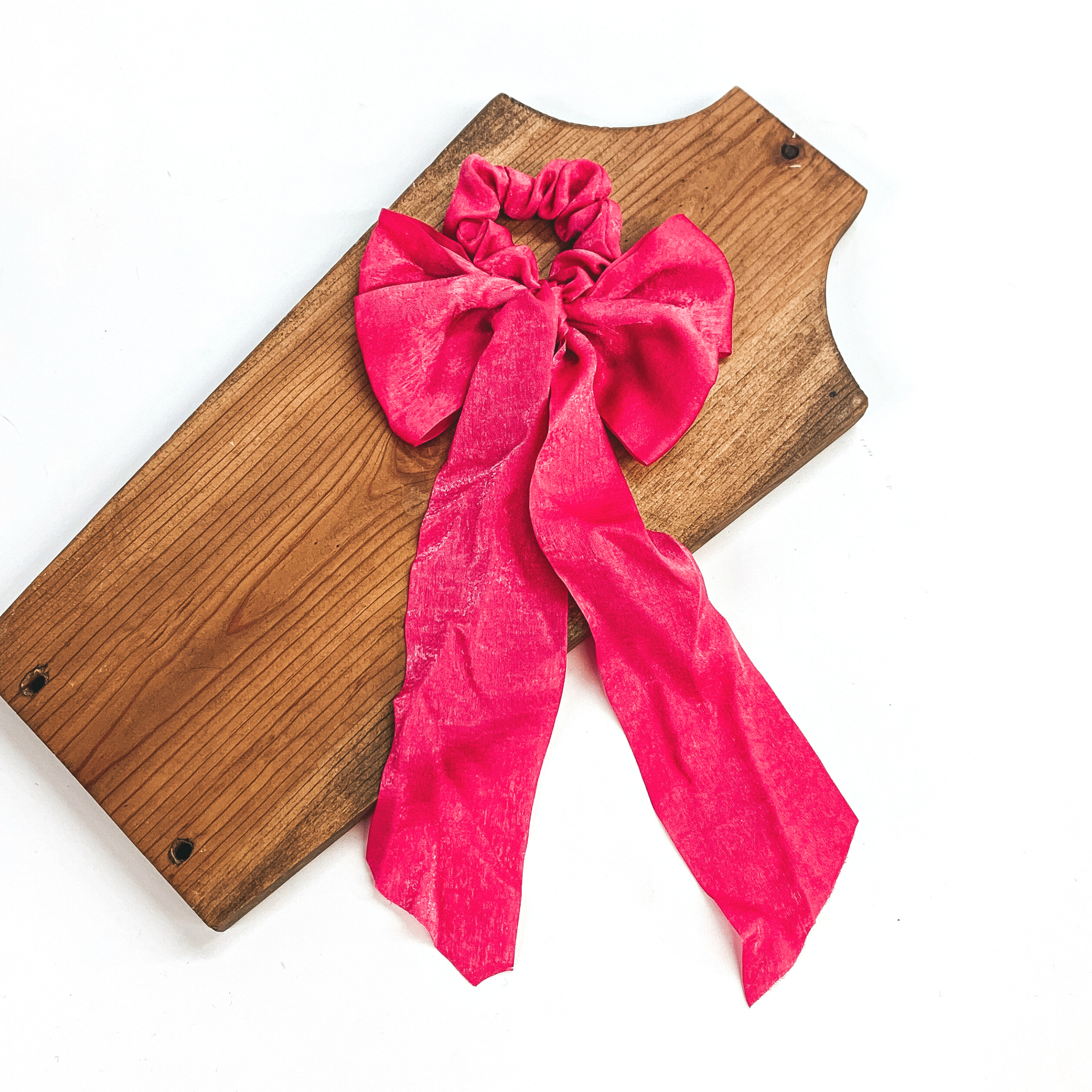 This is a hot pink satin scrunchie with a long bow, this scrunchie is taken on  a brown necklace board and on a white background.