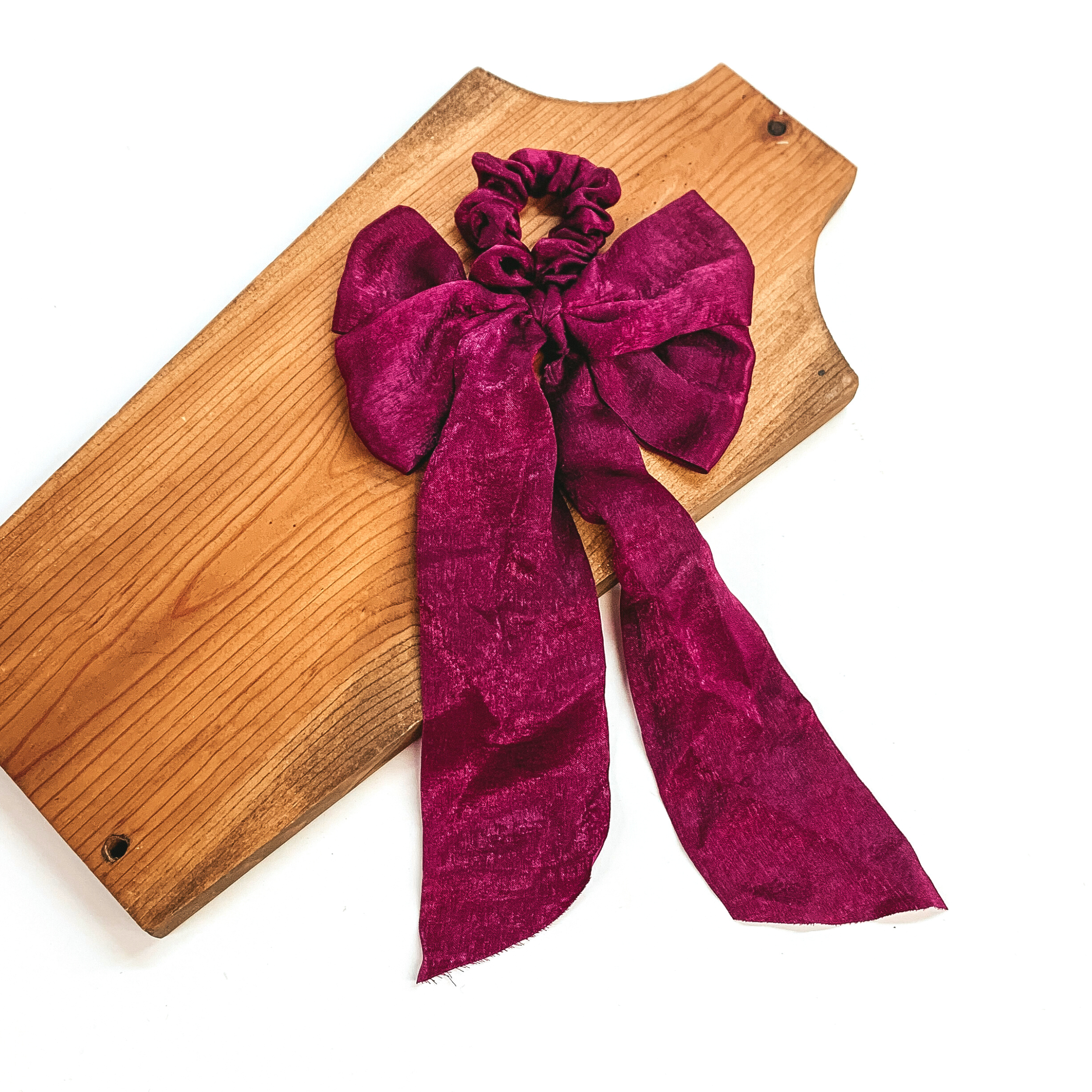 This is a maroon satin scrunchie with a long bow, this scrunchie is taken on  a brown necklace board and on a white background.