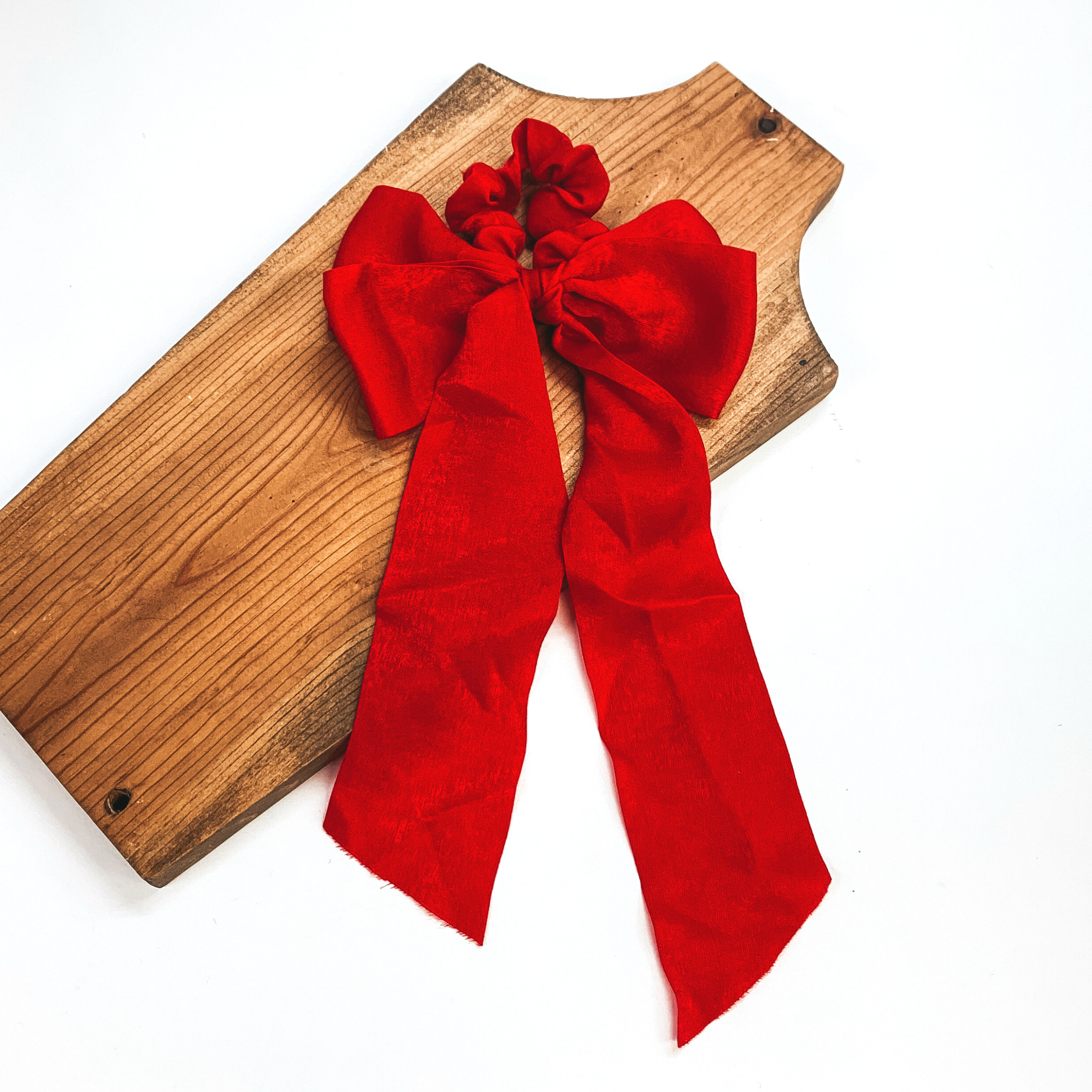This is a red satin scrunchie with a long bow, this scrunchie is taken on  a brown necklace board and on a white background.