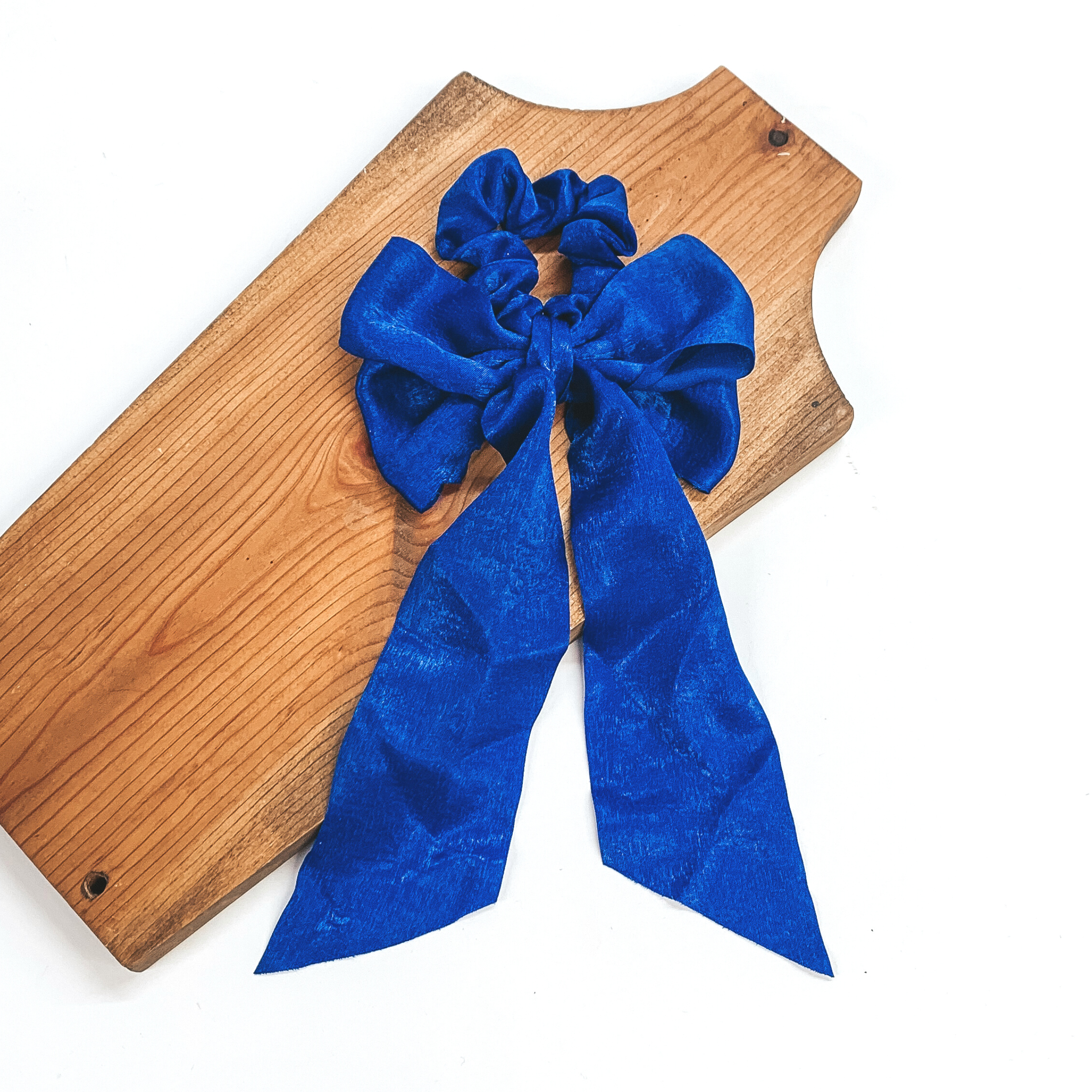 This is a royal blue satin scrunchie with a long bow, this scrunchie is taken on  a brown necklace board and on a white background.