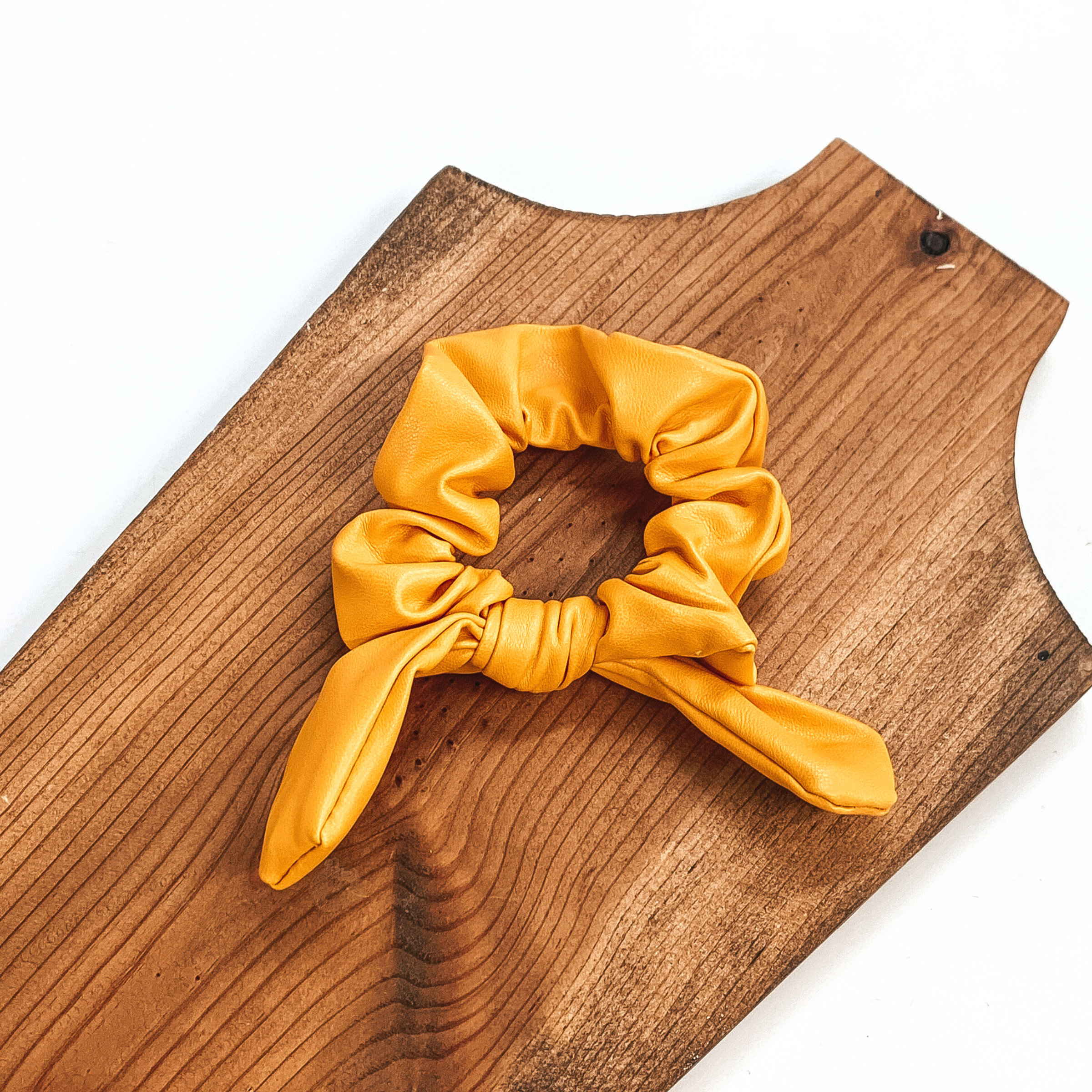 This is yellow faux leather scrunchie with a bow, this scrunchie is taken on  a brown necklace board and on a white background.