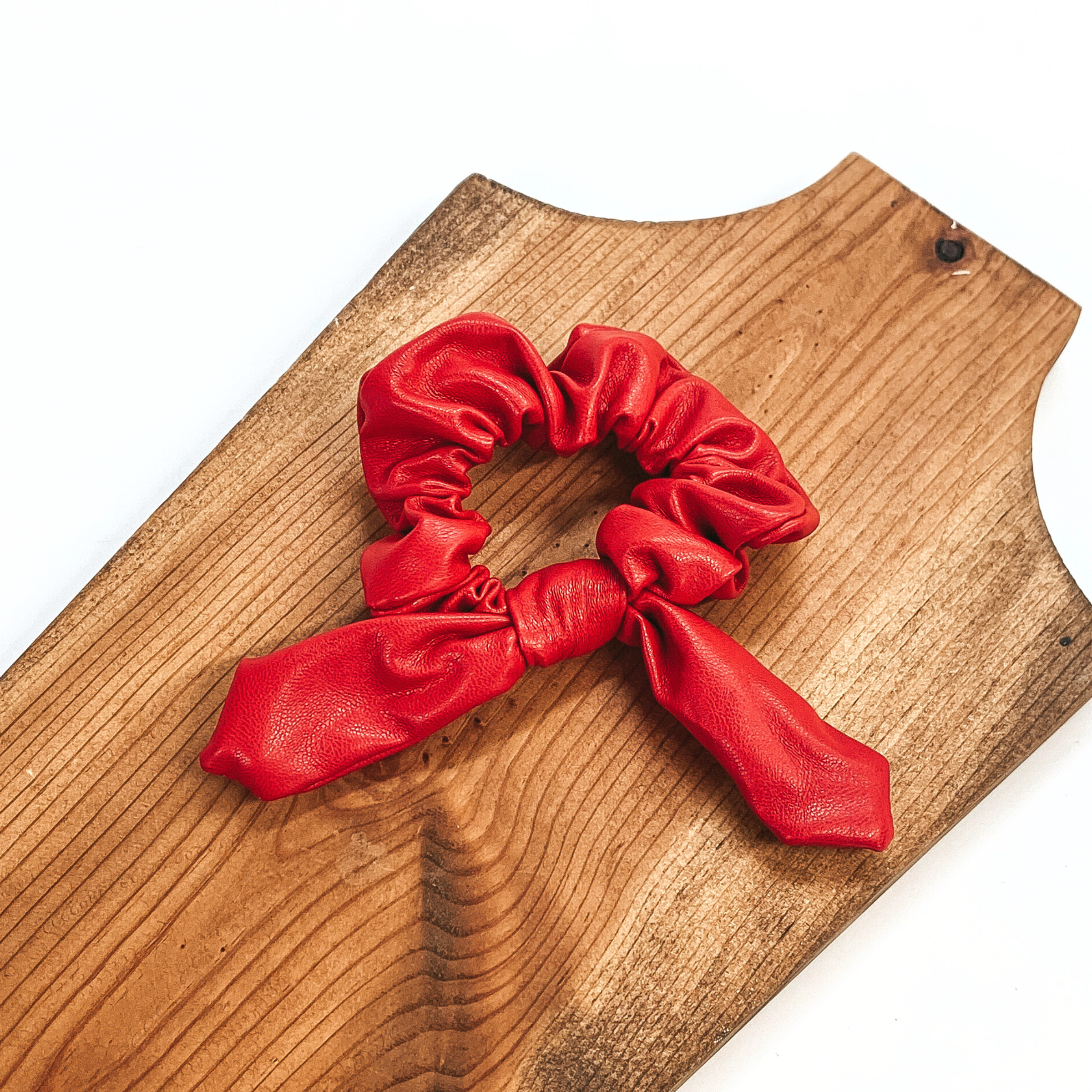 This is red faux leather scrunchie with a bow, this scrunchie is taken on  a brown necklace board and on a white background.
